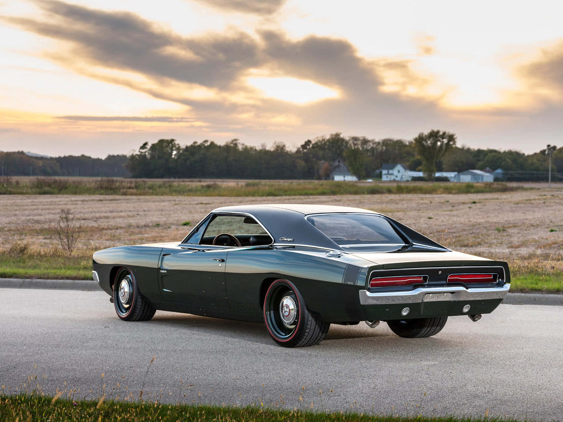 1969 年道奇 Charger 桌布 壁紙 — 一架時尚深綠色經典肌肉車停泊在日落時分的鄉村道路上，背景有金色雲彩和遠處的農舍。