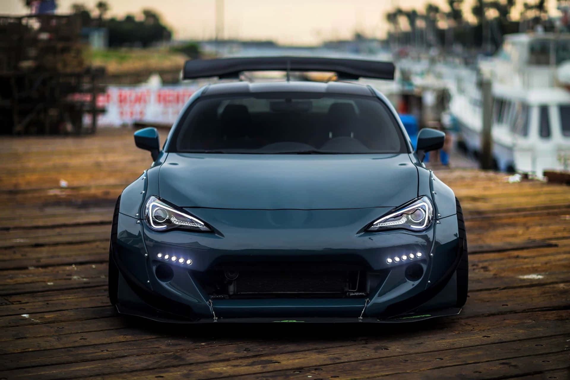 深藍綠色改裝跑車停泊在木碼頭上，背景有船隻，黃昏時分。 — Toyota Supra Mk4 壁紙