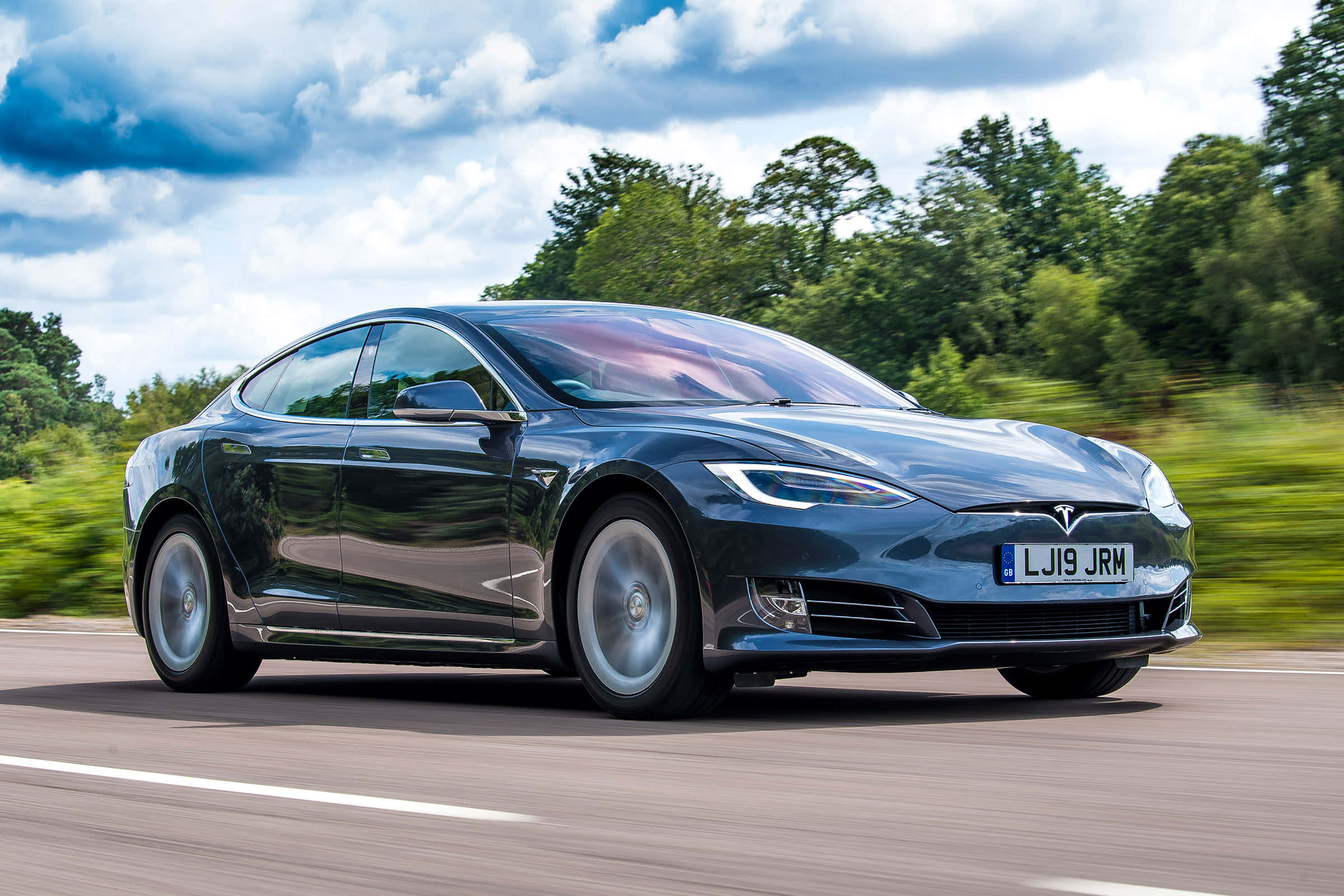特斯拉 桌布 壁紙 — 一架時尚深藍色Tesla Model S在陽光明媚的道路上行駛，背景是模糊的綠色樹木和戲劇性的多雲天空。