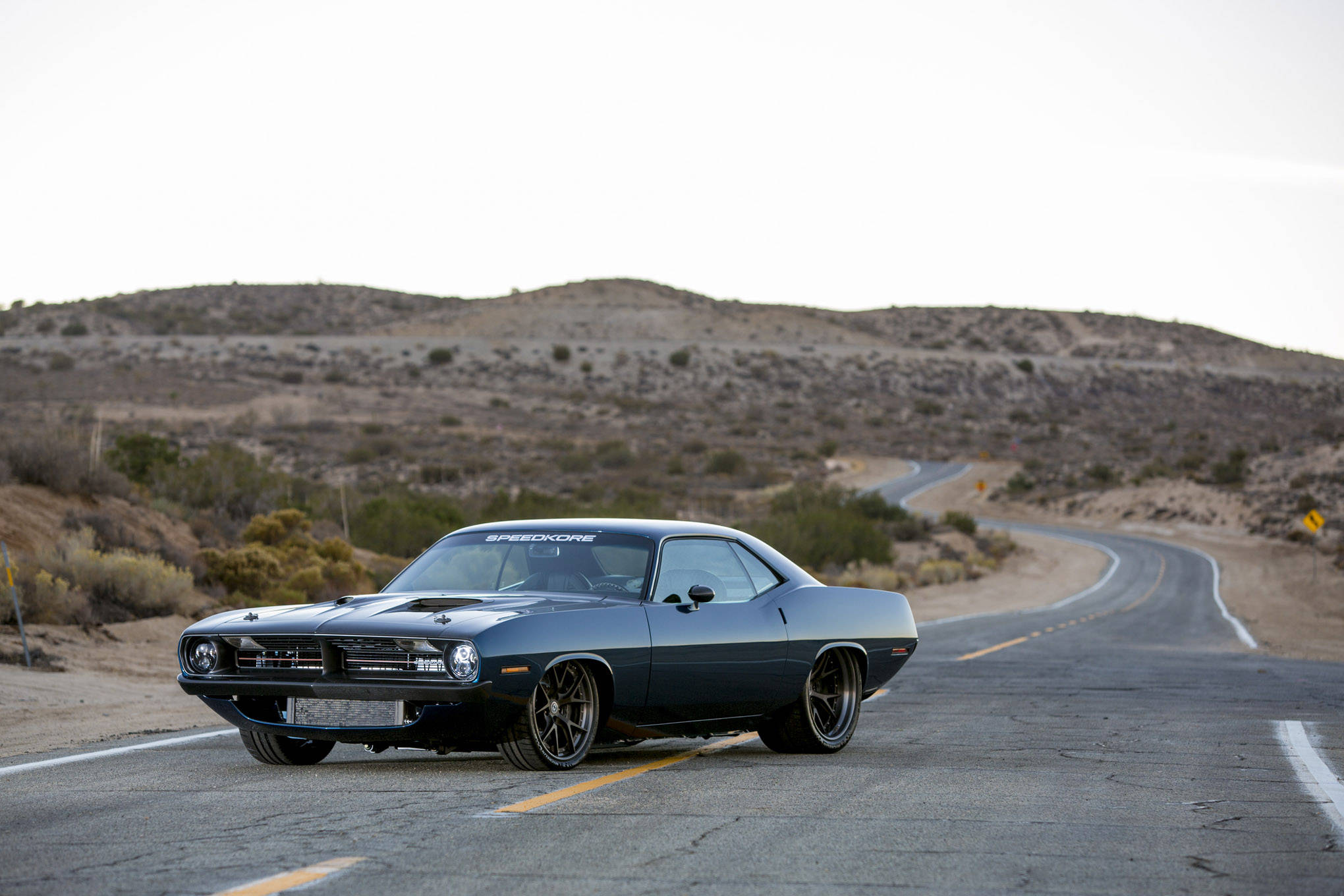 Vintage Car 桌布 壁紙 — 一架時尚深藍色經典肌肉車在蒼白天空下沿著蜿蜒的沙漠公路行駛。