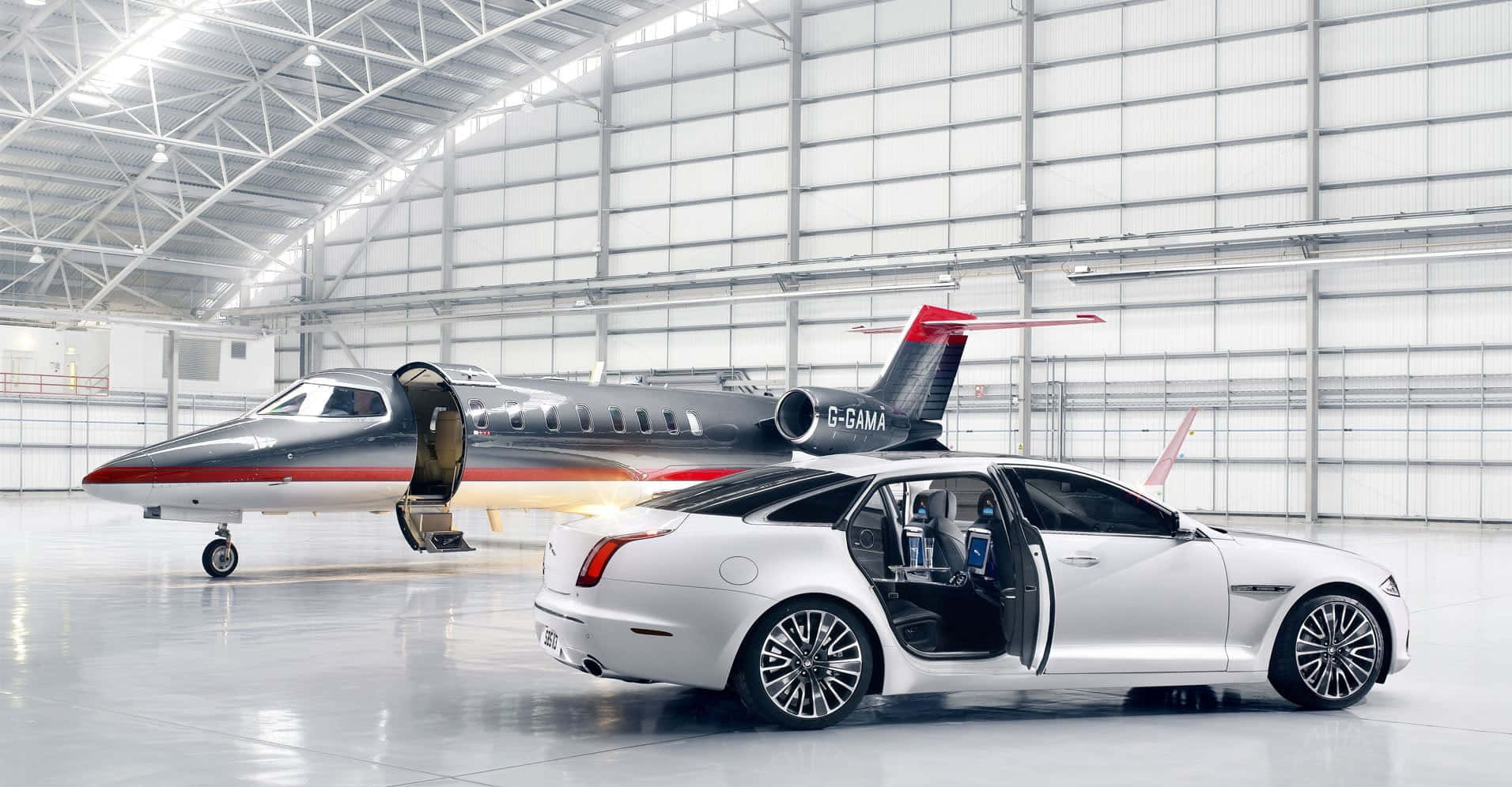 飛機 桌布 壁紙 — 一架時尚白色豪華轎車與私人飛機並排停放在寬敞、高天花板、地板拋光及工業照明的機庫內。