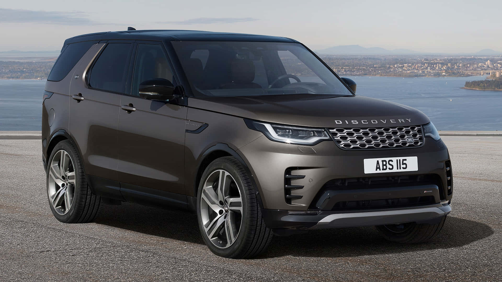 Land Rover Discovery 桌布 壁紙 — 一架時尚的啞光棕色 Land Rover Discovery 停泊在海岸觀景點，背景是城市景觀和水面。