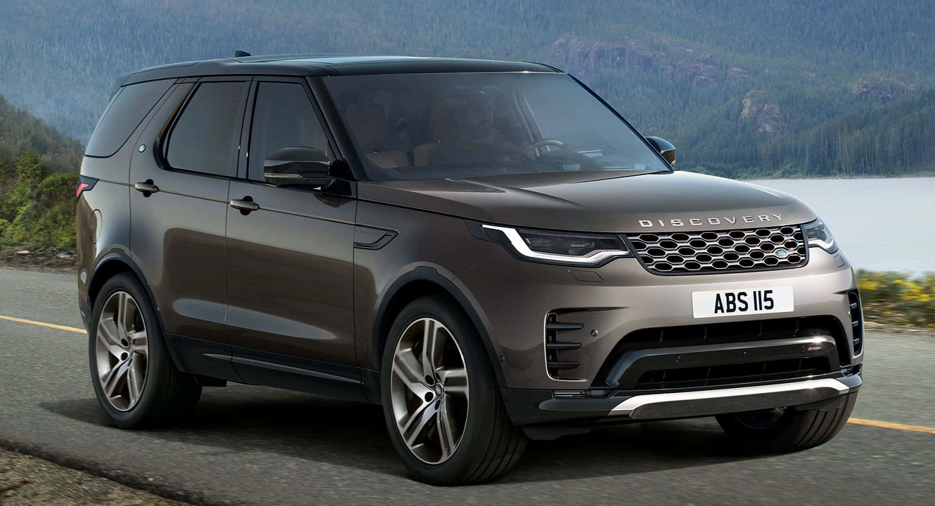 Land Rover Discovery 桌布 壁紙 — 一架時尚的啞光灰色 Land Rover Discovery 停在路邊，背景是森林覆蓋的山脈和湖泊。