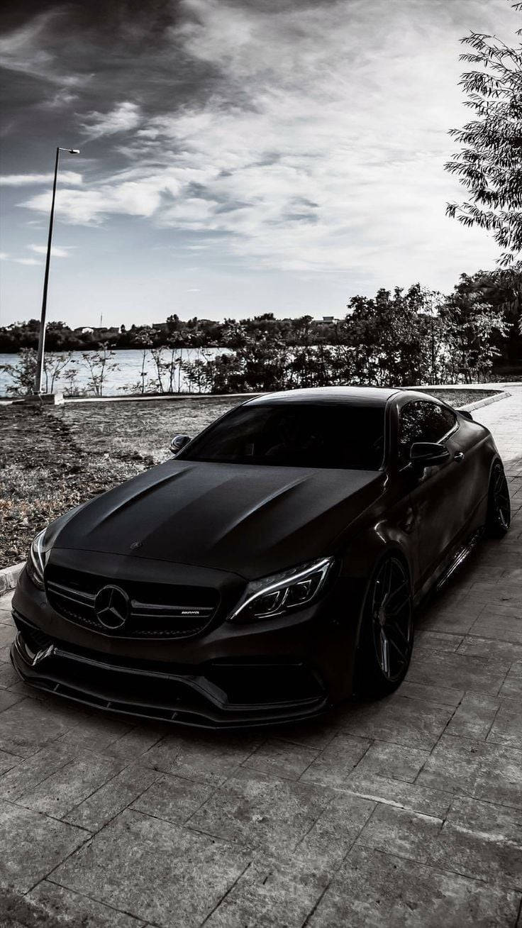 Amg 桌布 壁紙 — 一架時尚的啞光黑色Mercedes-AMG跑車停泊在平靜湖泊旁的石徑上，天空多雲。