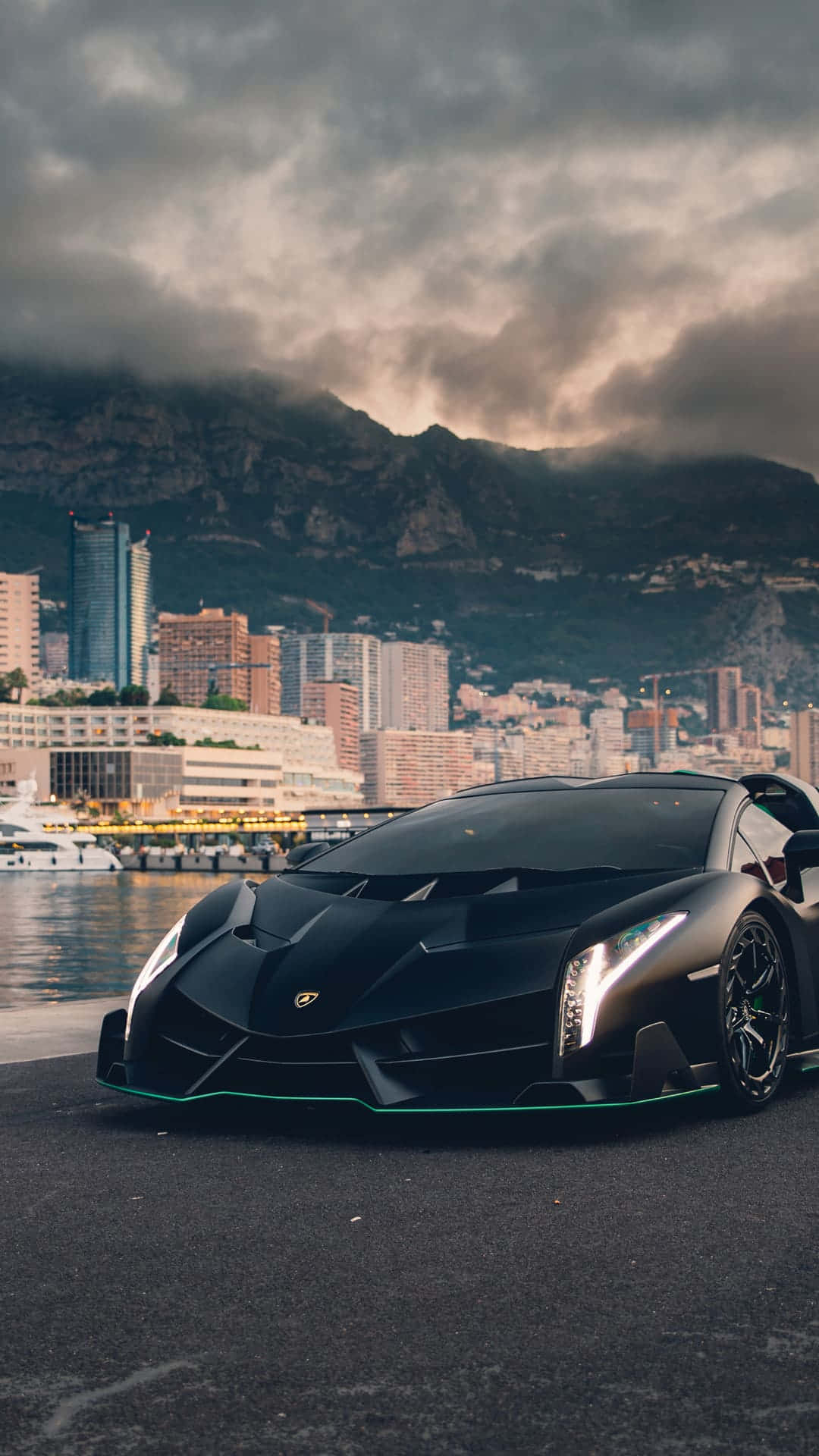 Pixel 3xl Lamborghini 桌布 壁紙 — 一架時尚的啞光黑色藍寶堅尼 Veneno 停泊在海濱碼頭上，背景是摩納哥的天際線和霧濛濛的山脈，天空風起雲湧。