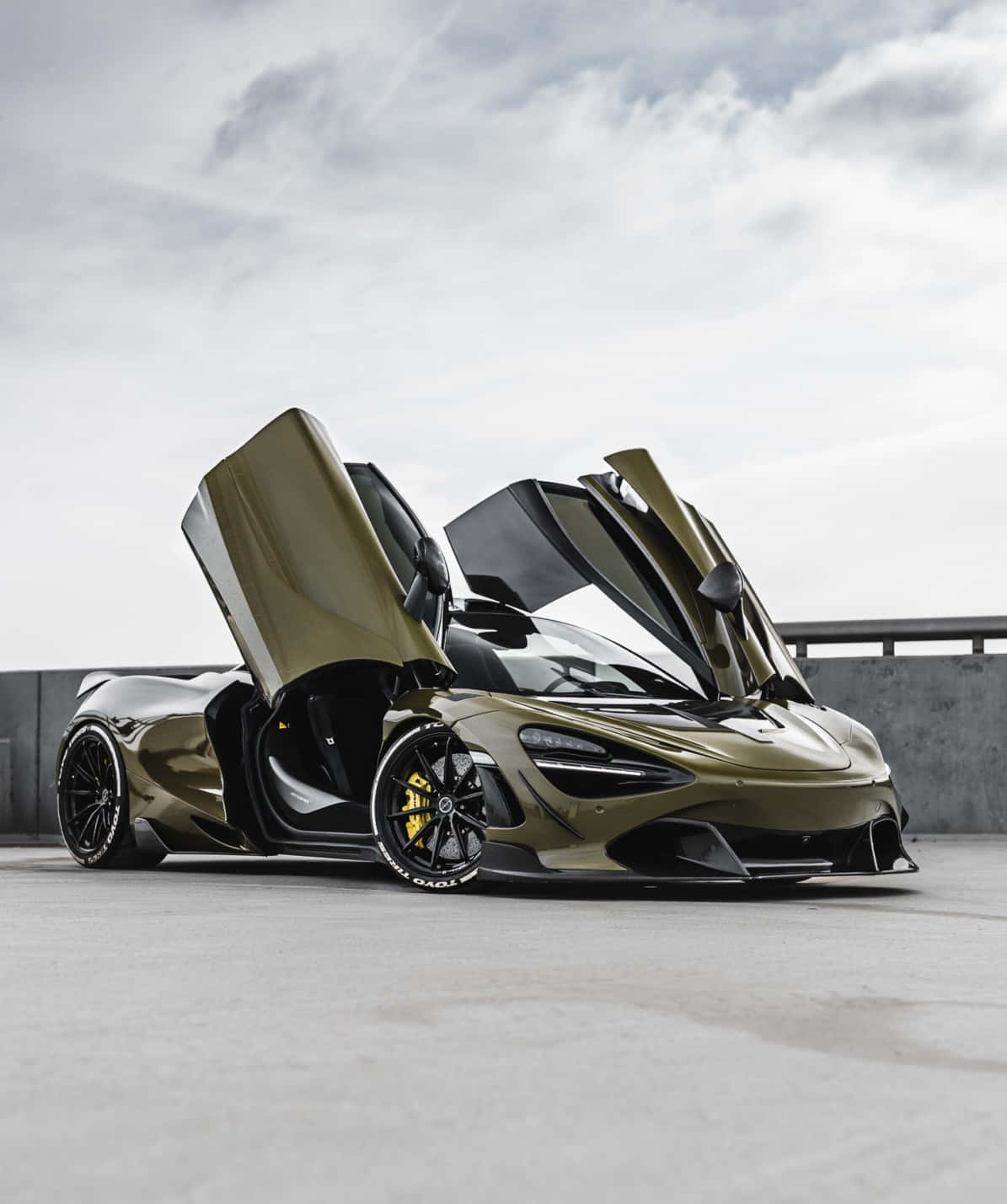 汽車 桌布 壁紙 — 一架時尚的橄欖綠色McLaren跑車，鷗翼車門打開，停放在混凝土平台上，天空多雲。