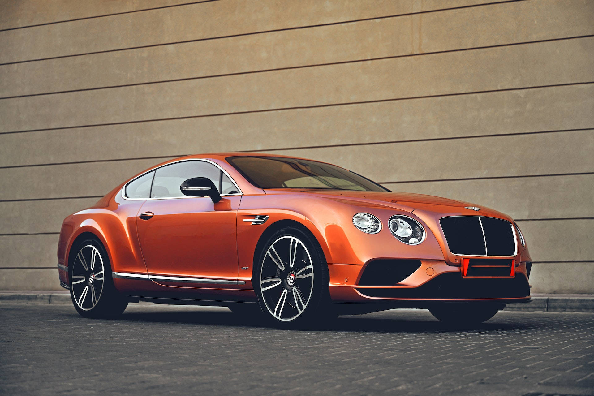 bentley HD 桌布 壁紙 — 一架時尚的橙色 Bentley Continental GT 轎跑車停靠在米色牆壁旁的鋪砌地面上。