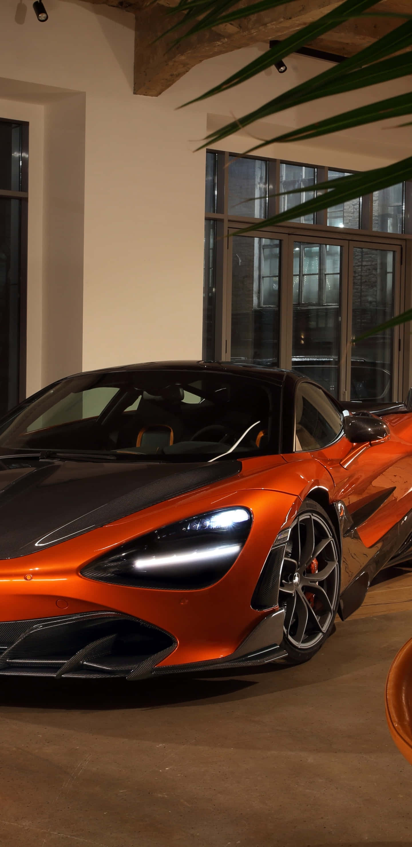 橘色 桌布 壁紙 — 一架時尚的橙色McLaren跑車在溫暖的室內燈光下，在大窗戶和綠色棕櫚葉旁閃閃發光。