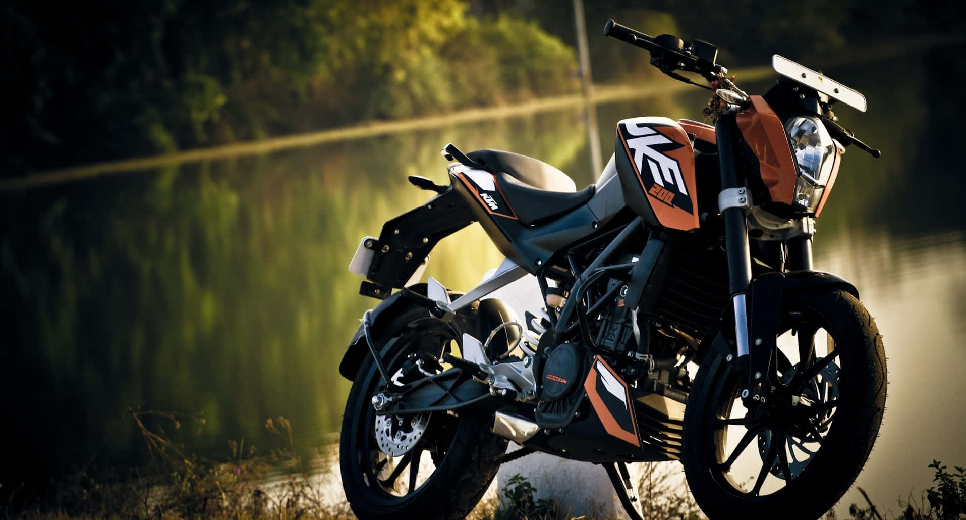 HD Bikes 桌布 壁紙 — 一架時尚的橙色和黑色KTM機車在黃金時刻停泊在霧氣瀰漫的湖邊，背景是茂密的綠樹。