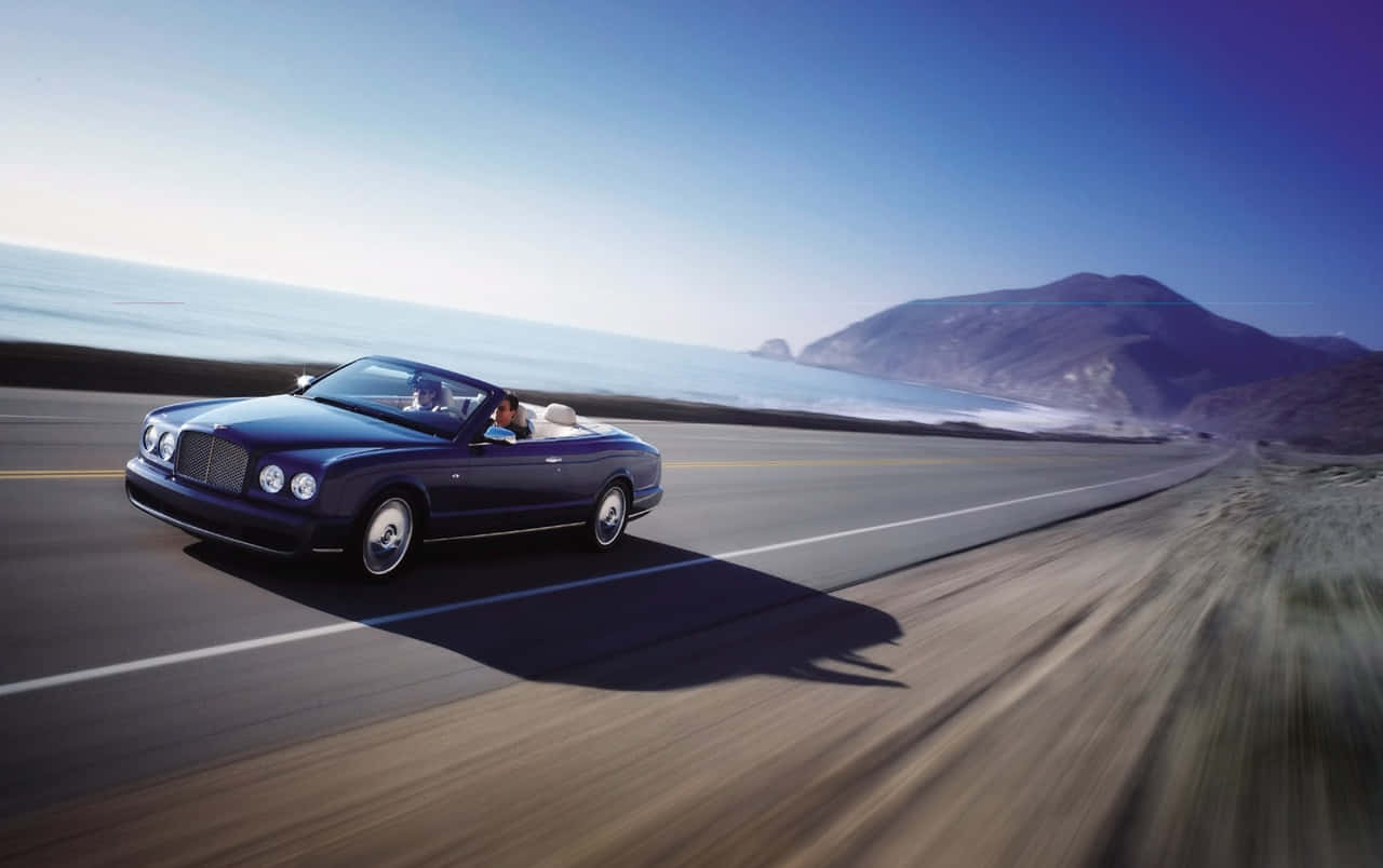 Bentley Azure 桌布 壁紙 — 一架時尚的海軍藍色敞篷跑車沿著海岸公路飛馳，背景是海洋和山脈，天空清澈湛藍。