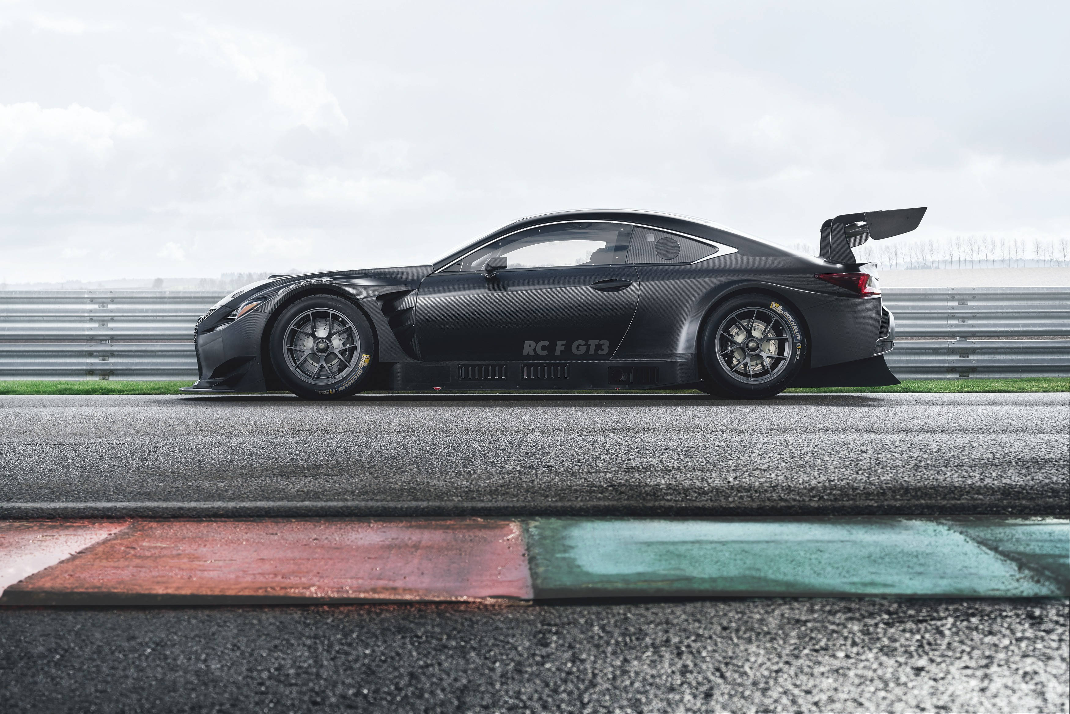 lexus iPhone 桌布 壁紙 — 一架時尚的消光黑色 Lexus RC F GT3 賽車，在陰天之下停放在濕滑的賽道上，旁邊有紅色和綠色的路緣石。