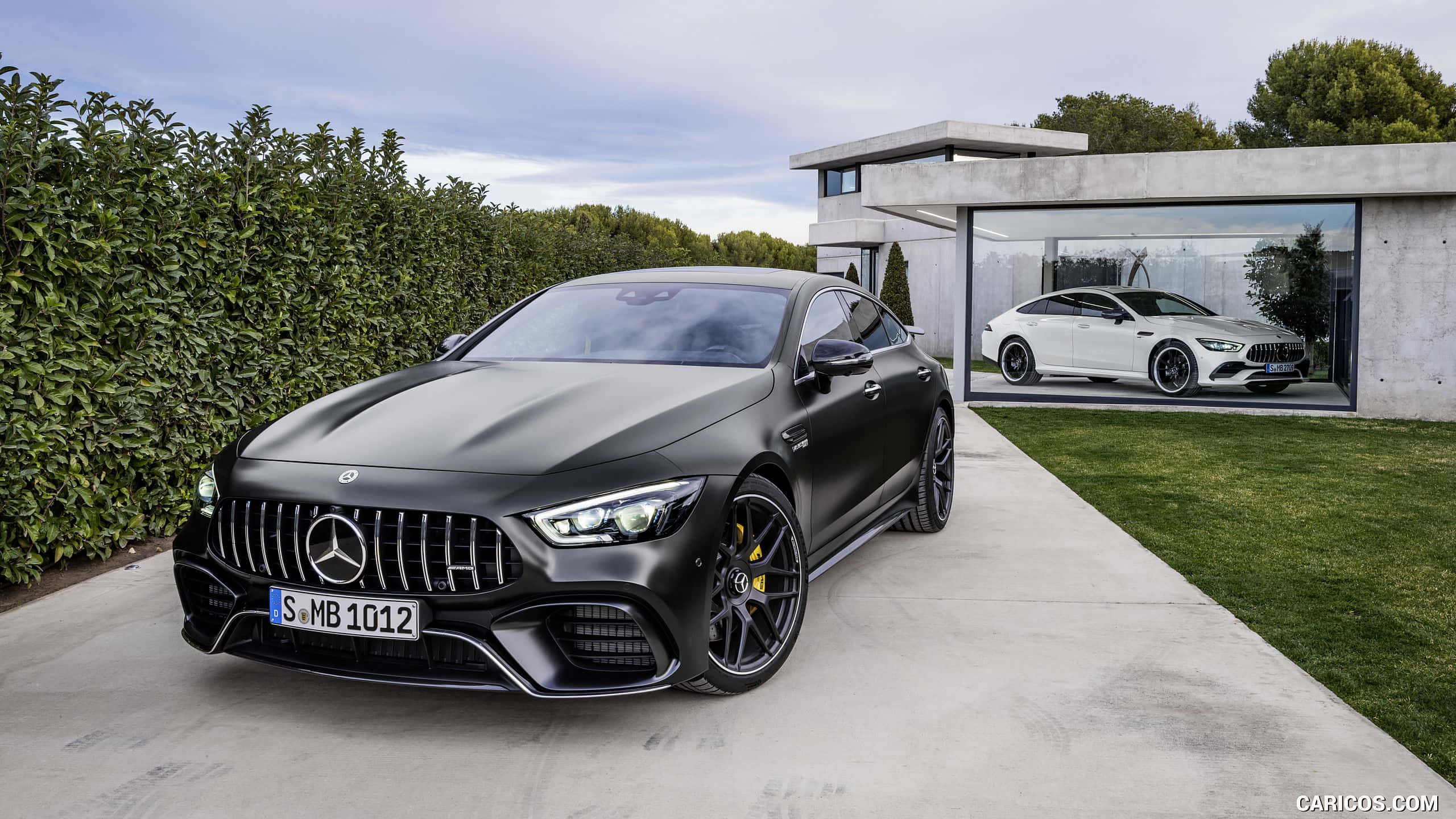Mercedes 桌布 壁紙 — 一架時尚的消光黑色Mercedes-AMG GT停放在混凝土車道上，旁邊是現代玻璃車庫，裡面露出一輛白色AMG。