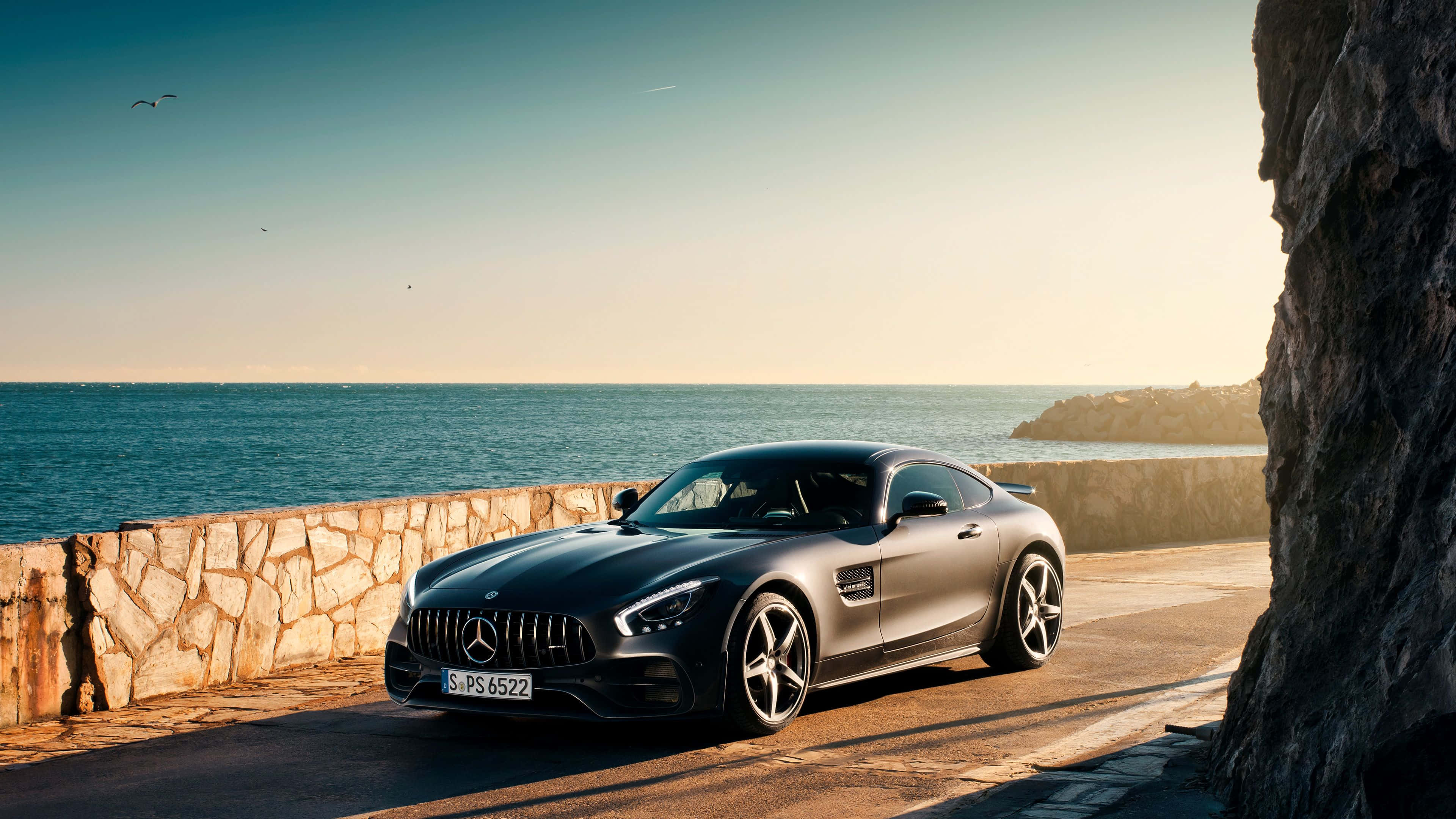 4K 奔馳壁紙 桌布 壁紙 — 一架時尚的消光黑色Mercedes-AMG GT跑車停泊在沿海道路上，旁邊是石牆，背景是日落時分的海洋和懸崖。
