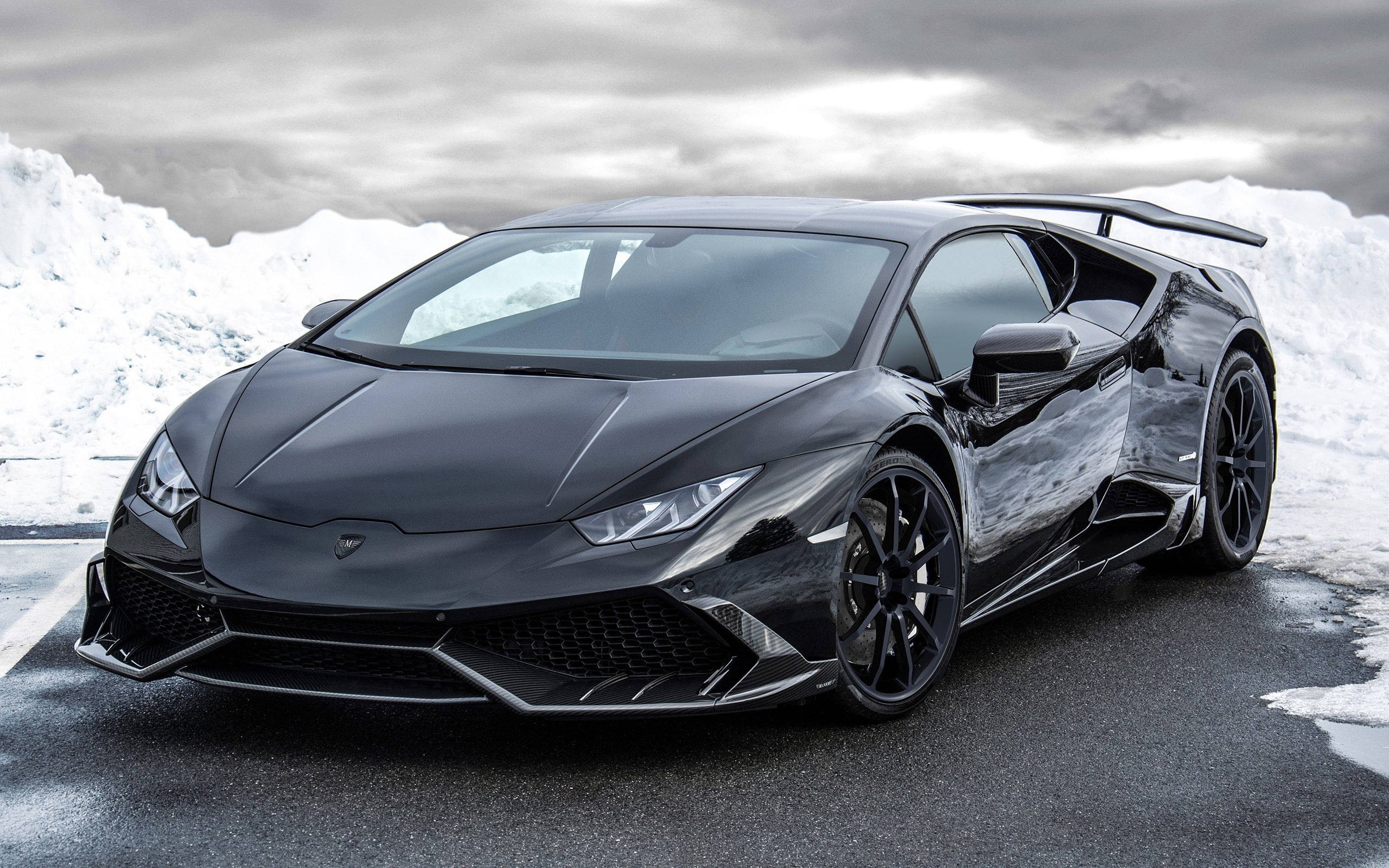 Lamborghini 4K 桌布 壁紙 — 一架時尚的消光黑色藍寶堅尼Huracán跑車停放在濕漉漉的柏油路上，背景是雲層密布的天空下積雪覆蓋的山脈。