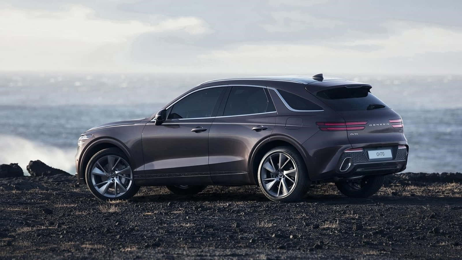 Genesis Gv70 桌布 壁紙 — 一架時尚的深灰色 Genesis GV70 SUV 停放在黑色火山岩上，旁邊是波濤洶湧的海洋，天空多雲。
