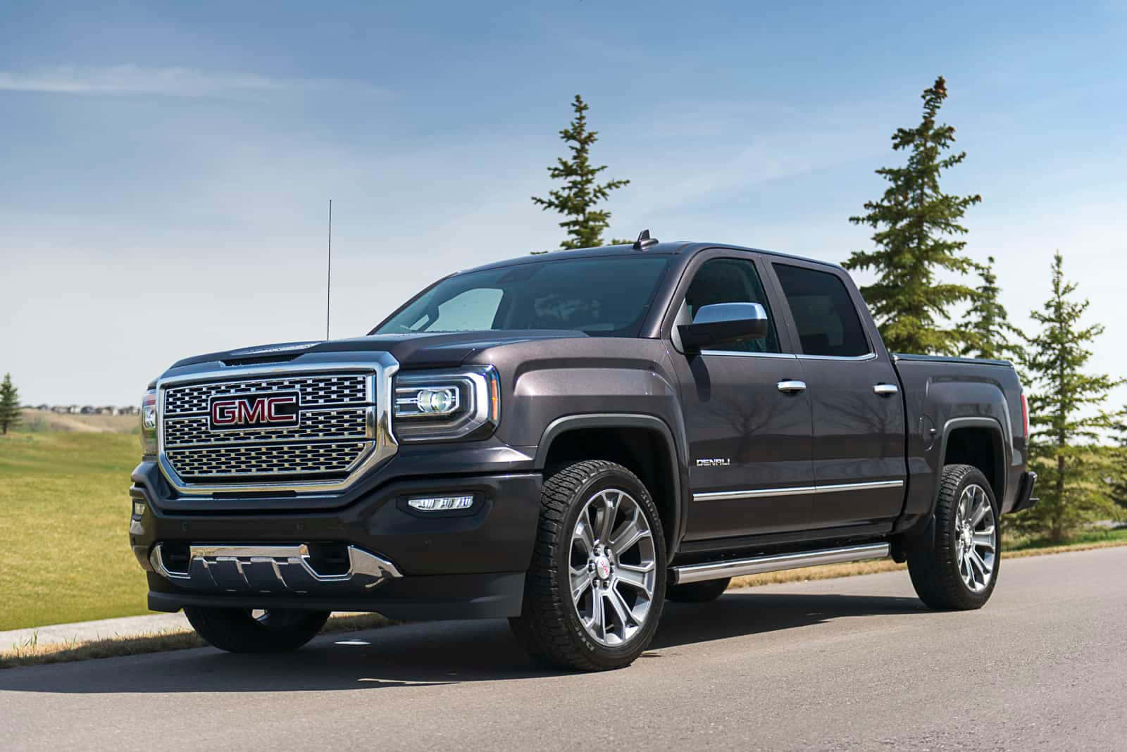 GMC 桌布 壁紙 — 一架時尚的深灰色 GMC Sierra Denali 貨卡停在鋪砌的道路上，周圍有常綠樹和草地，天空清澈湛藍。
