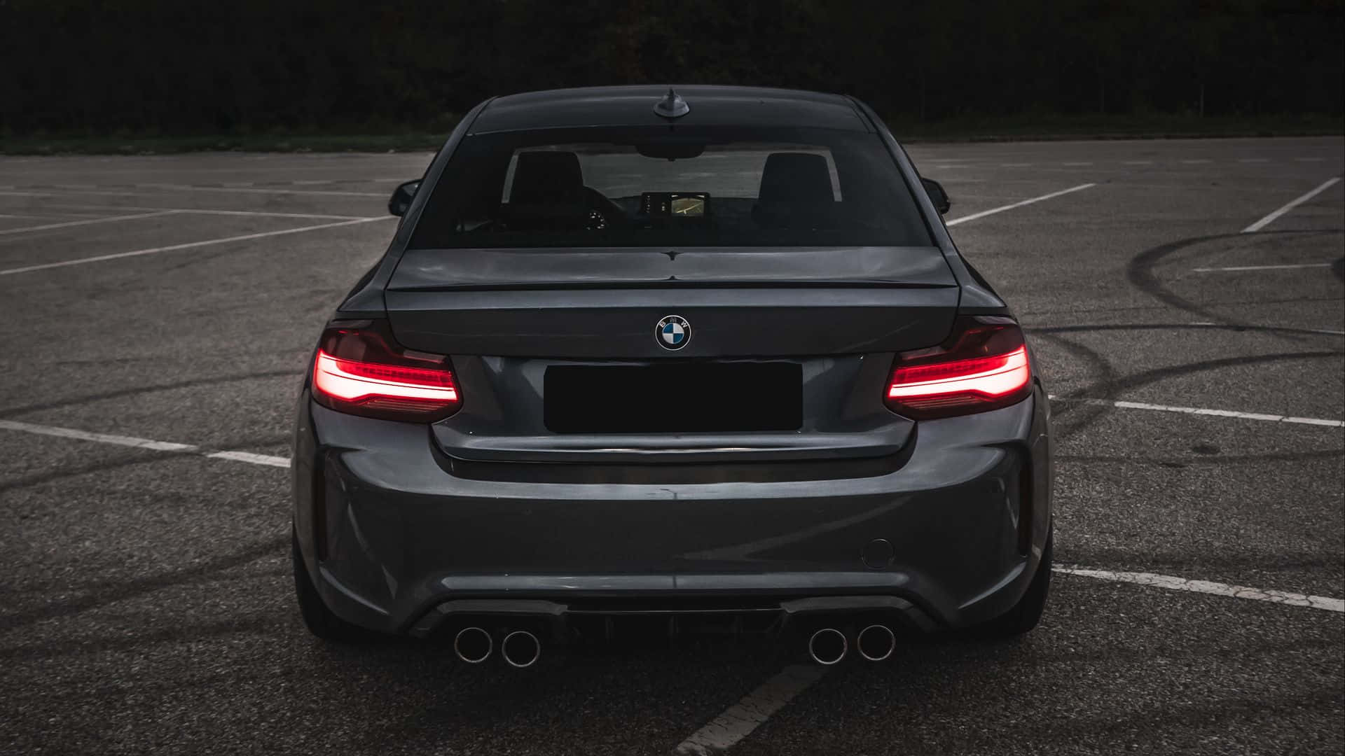Bmw M Series 桌布 壁紙 — 一架時尚的深灰色BMW M2跑車，帶有發光的紅色尾燈，停泊在黃昏時空無一人的瀝青停車場。