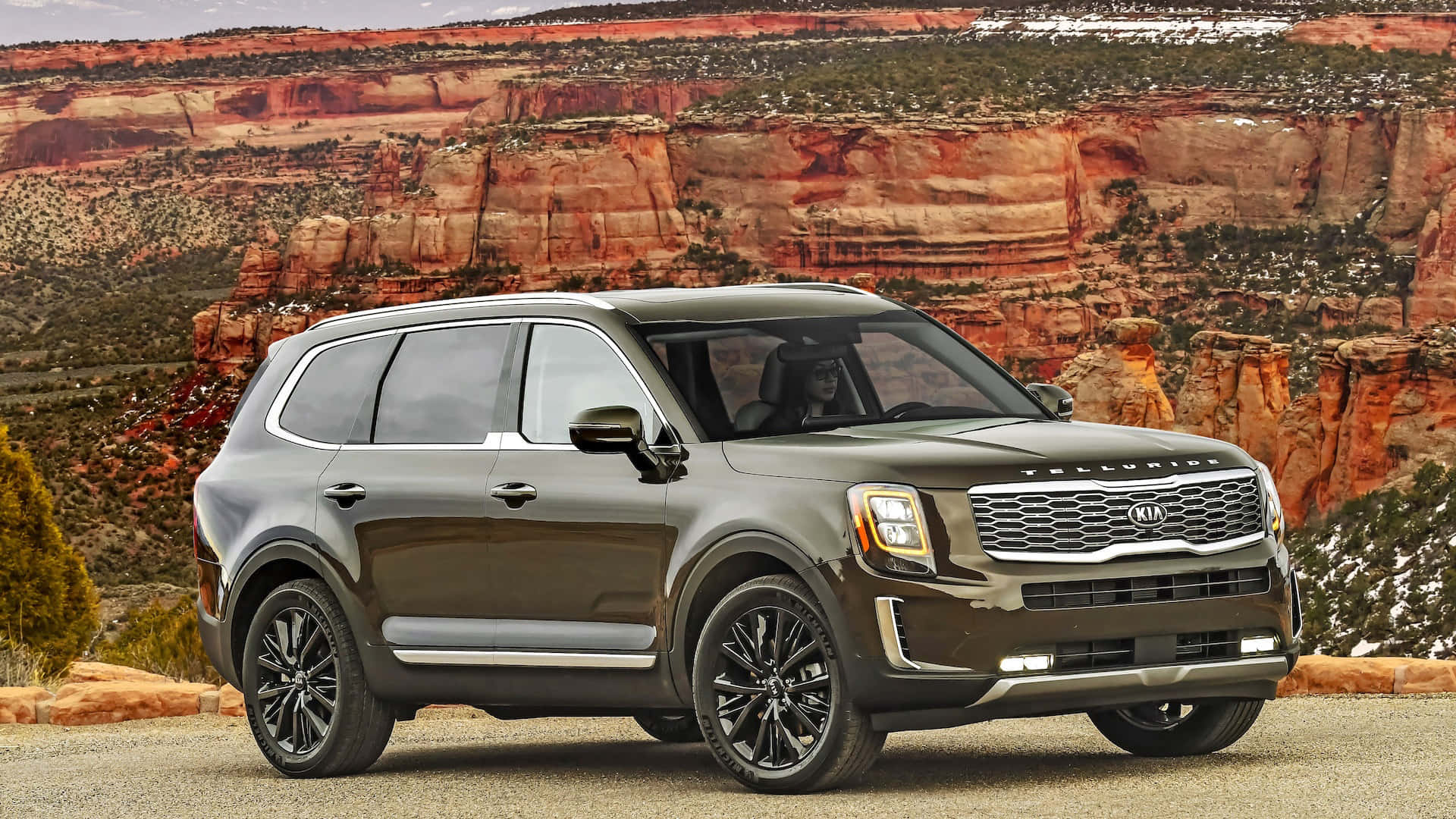 Kia Telluride 桌布 壁紙 — 一架時尚的深灰色Kia Telluride SUV停泊在碎石路上，背景是戲劇性的紅色岩石峽谷懸崖。