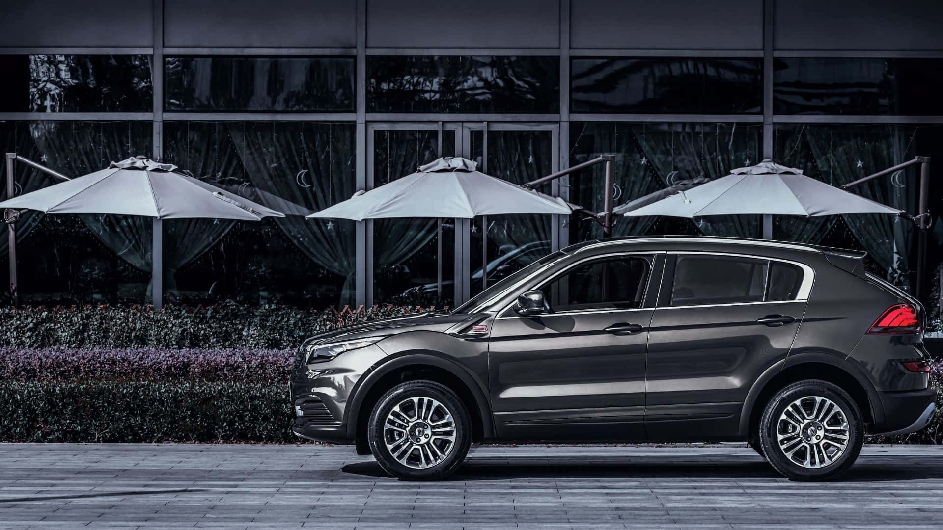 Qoros 桌布 壁紙 — 一架時尚的深灰色SUV停放在瓷磚庭院上，旁邊有白色遮陽傘和現代玻璃建築。