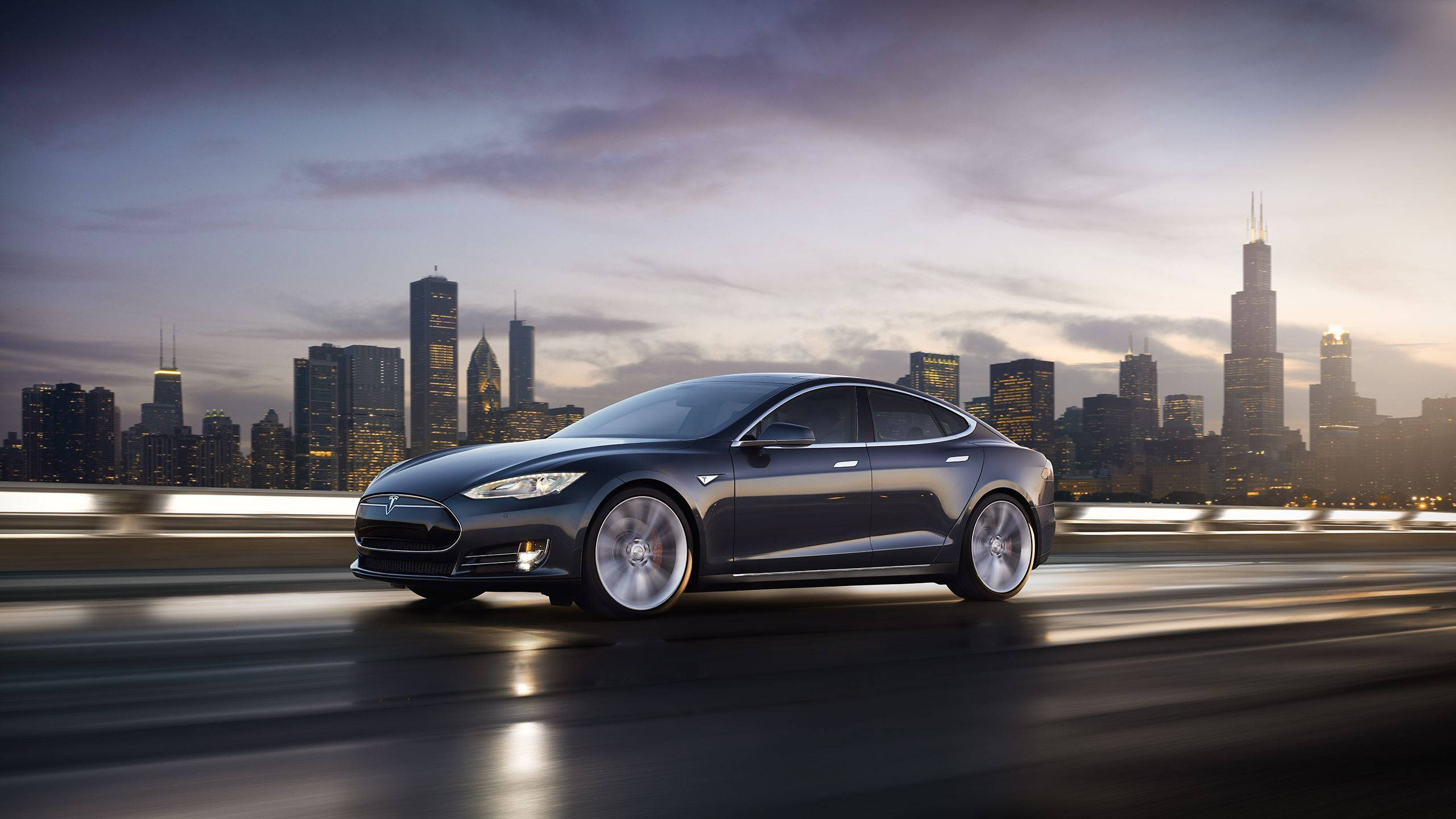 特斯拉 桌布 壁紙 — 一架時尚的深灰色Tesla Model S在高速公路上巡航，芝加哥璀璨的天際線在黃昏時分在它背後閃耀。