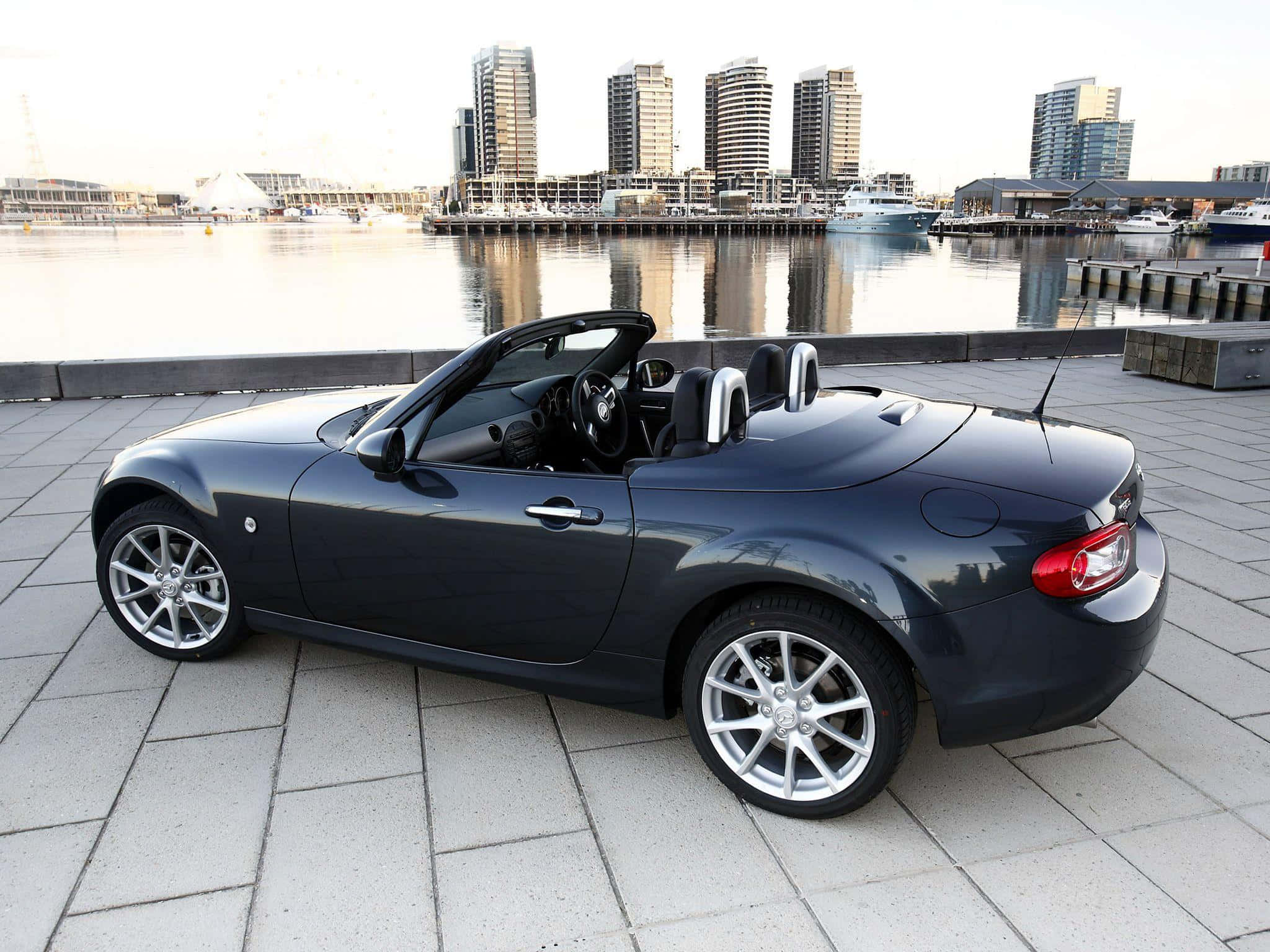 Miata 桌布 壁紙 — 一架時尚的深灰色敞篷跑車停泊在鋪砌的海濱長廊上，背後現代化的摩天大樓倒映在平靜的水面上。