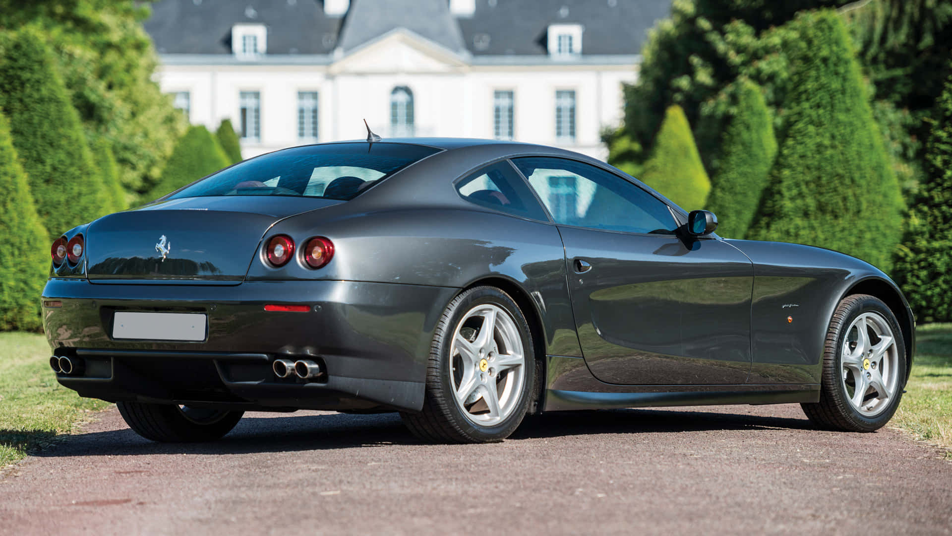 Ferrari 612 Scaglietti 桌布 壁紙 — 一架時尚的深灰色法拉利跑車停放在碎石車道上，背景是一座宏偉的白色豪宅和修剪整齊的綠色樹籬。