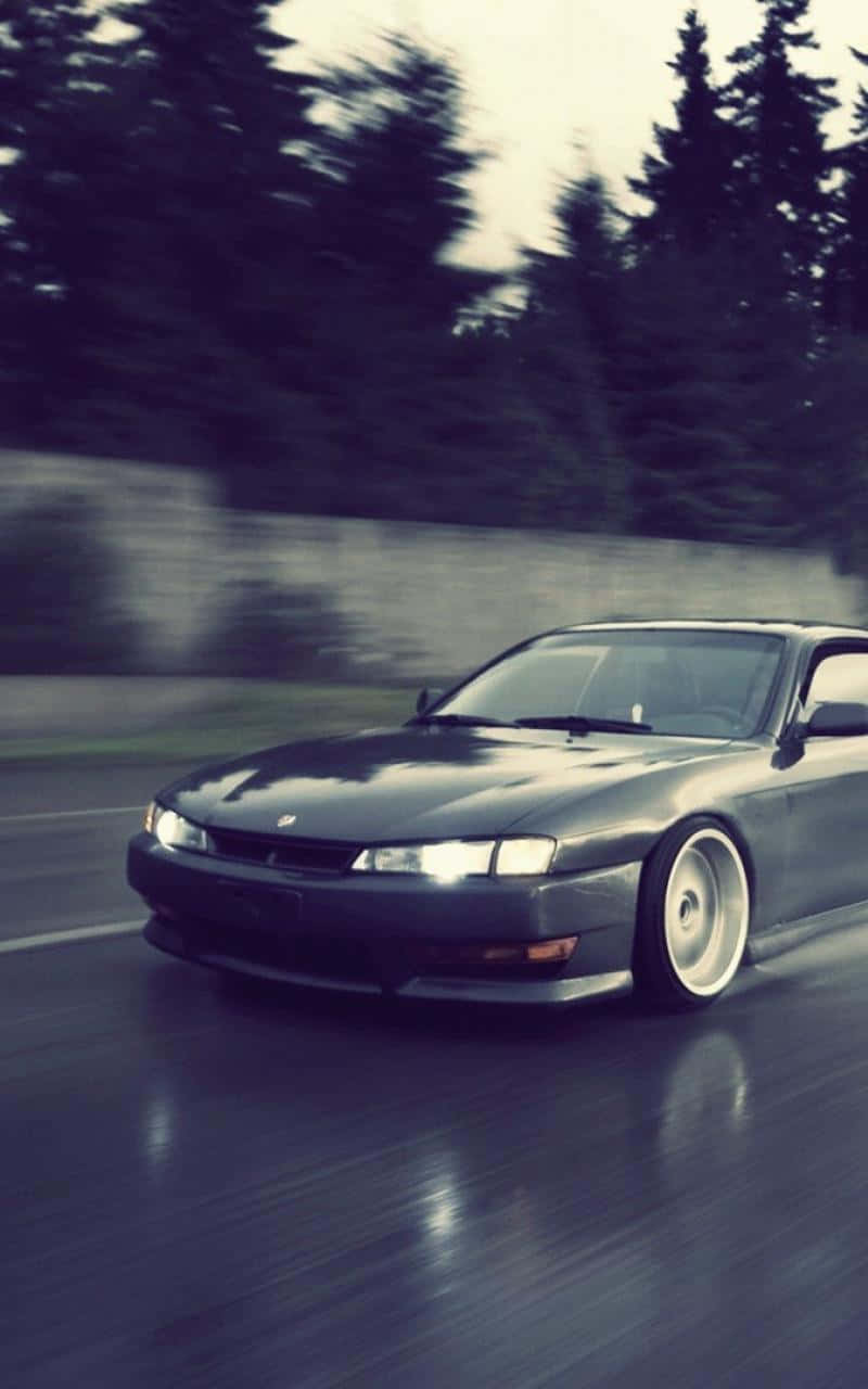 JDM 桌布 壁紙 — 一架時尚的深灰色跑車在濕滑的道路上飛馳，背景是模糊的樹木。