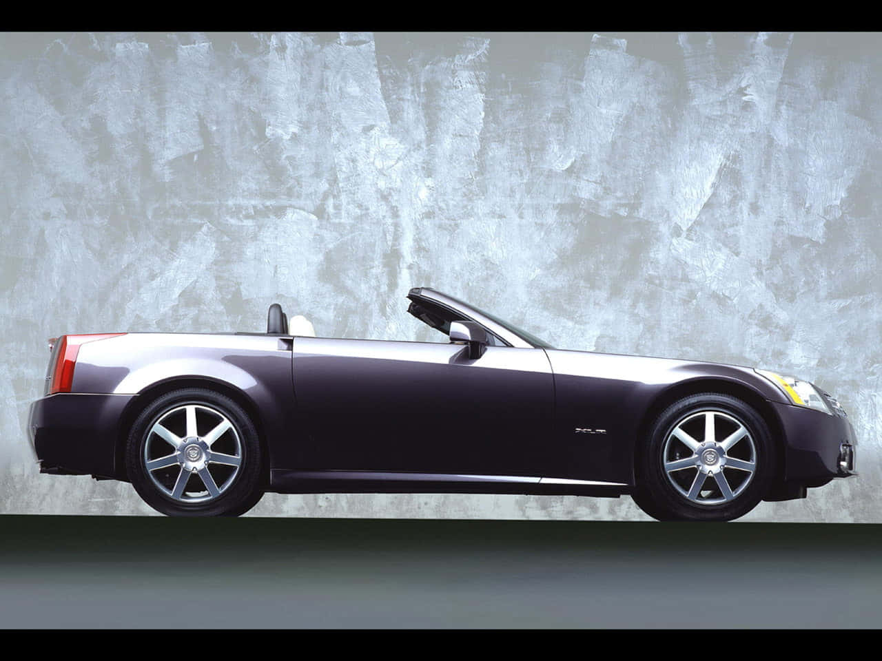 Cadillac Xlr 桌布 壁紙 — 一架時尚的深紫色敞篷跑車，帶有銀色飾邊和鉻合金輪圈，側面立於有紋理的灰色混凝土牆前。