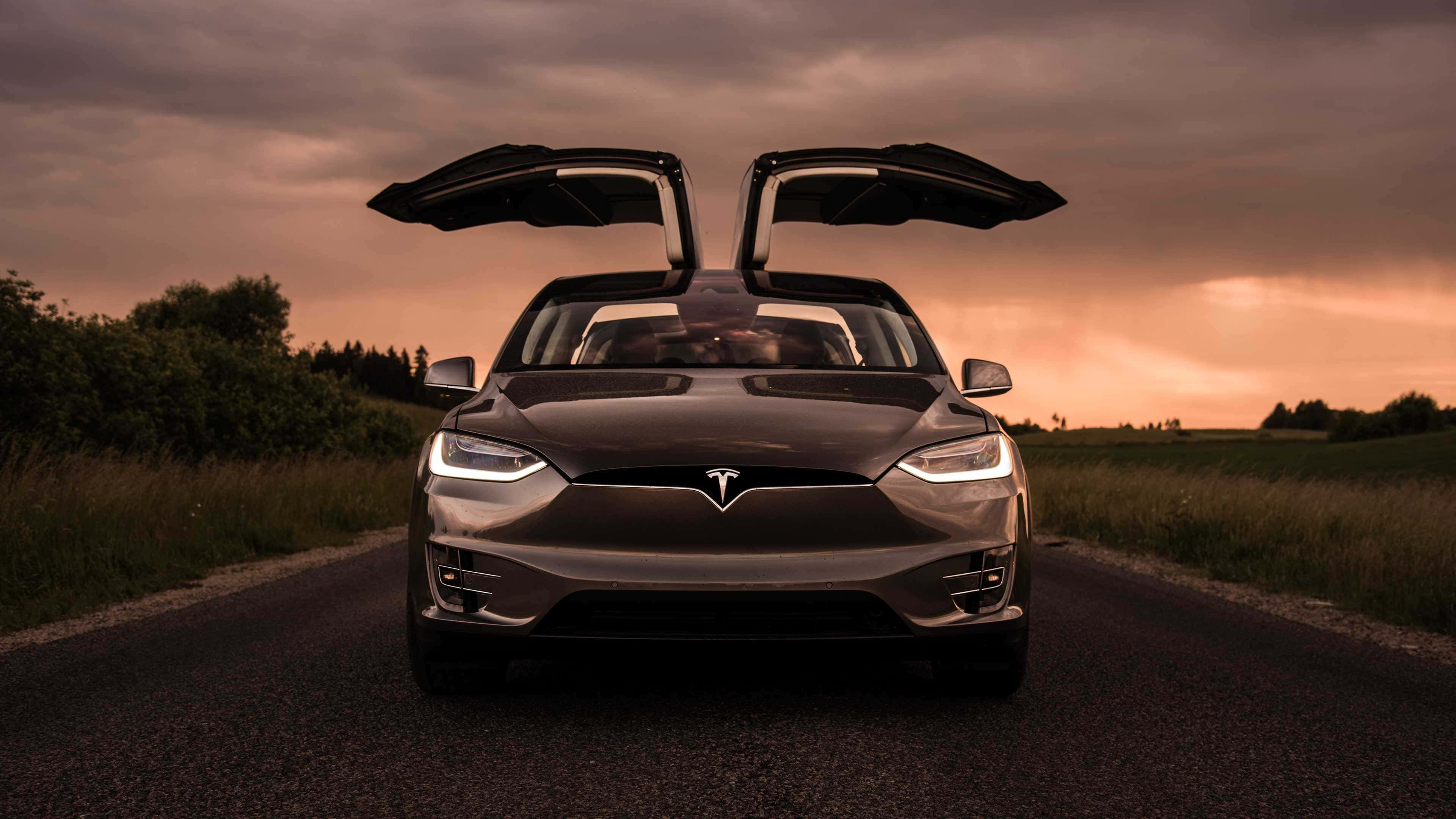 特斯拉 桌布 壁紙 — 一架時尚的深色Tesla，打開鷹翼門，停在鄉村道路上，背景是戲劇性的日落天空。