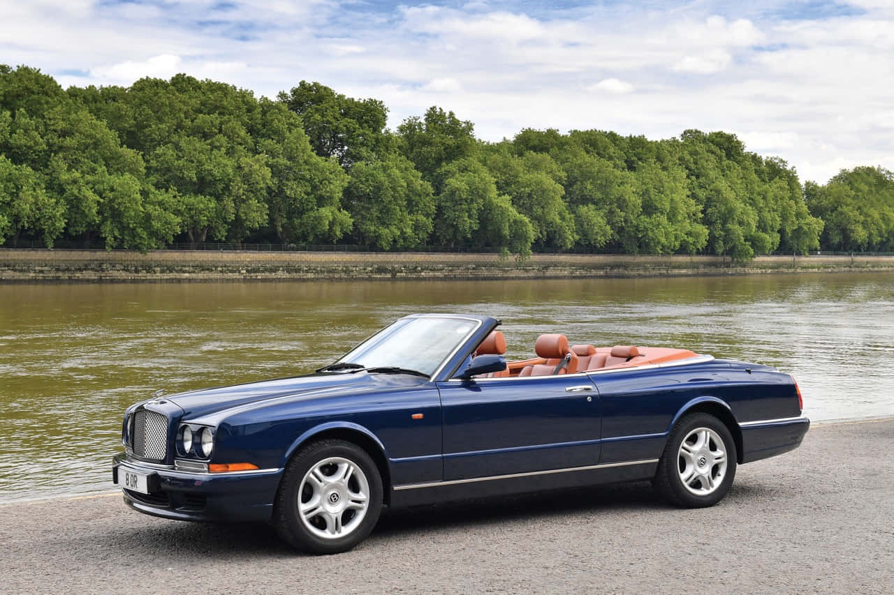 Bentley Azure 桌布 壁紙 — 一架時尚的深藍色賓利敞篷車，配橙色皮革座椅，停泊在平靜的河邊，兩旁綠樹成蔭，天空局部多雲。