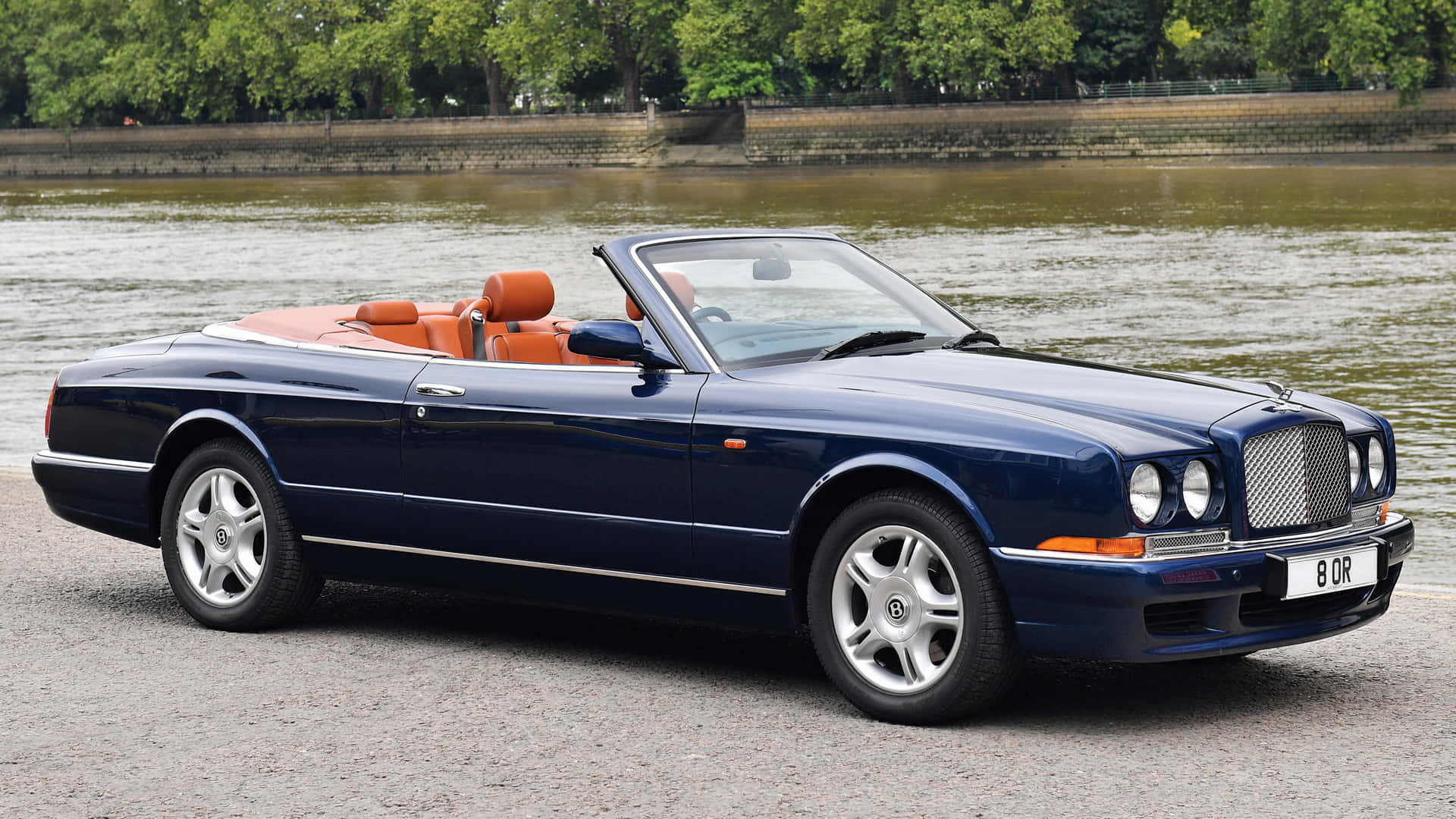 Bentley Azure 桌布 壁紙 — 一架時尚的深藍色賓利敞篷車，配橙色皮革座椅，在日間停泊在河岸邊。