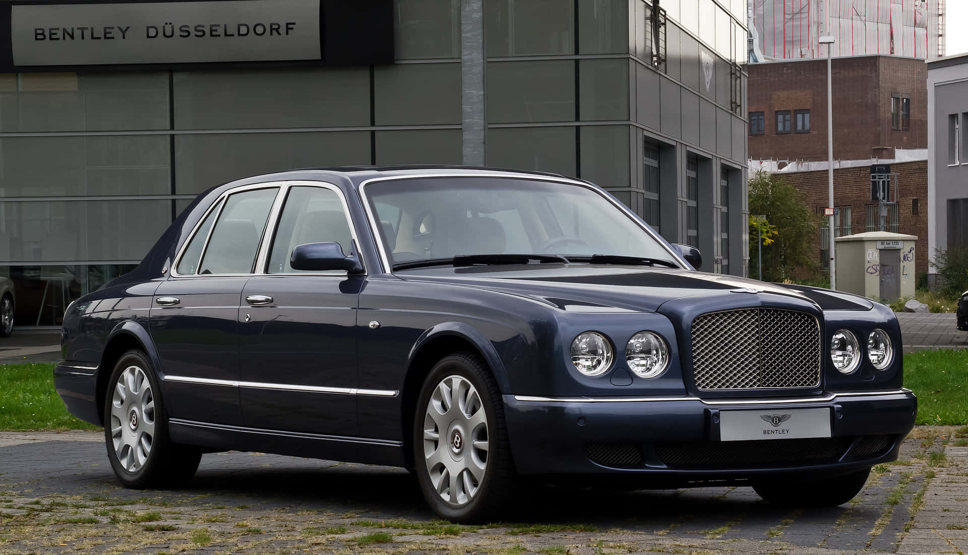 Bentley Arnage 桌布 壁紙 — 一架時尚的深藍色賓利轎車停泊在鵝卵石路上，前面是現代玻璃建築，帶有'BENTLEY DÜSSELDORF'標誌。