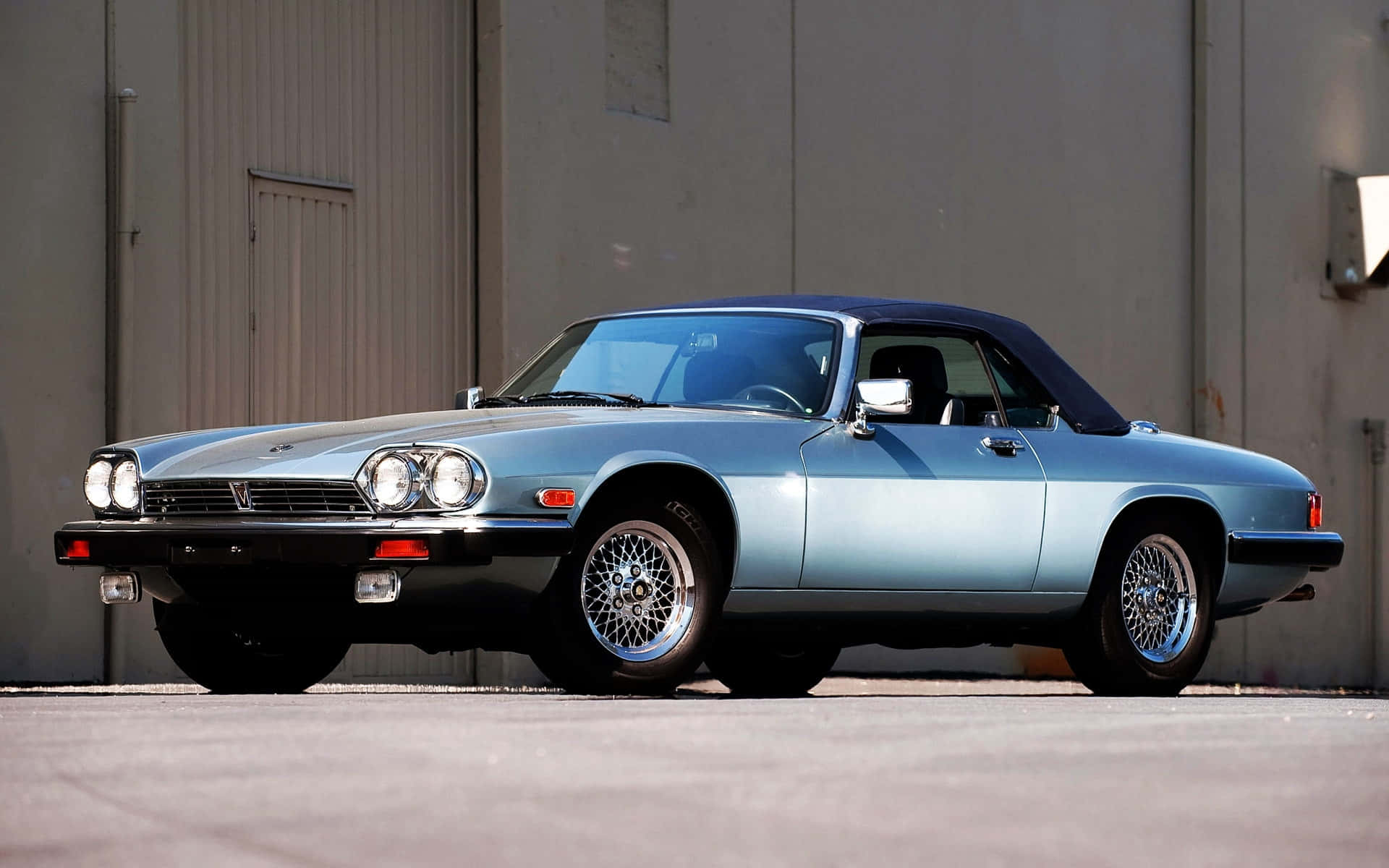 Jaguar Xjs 桌布 壁紙 — 一架時尚的淺藍色經典敞篷跑車，配備黑色軟頂，停放在樸素的工業建築前。