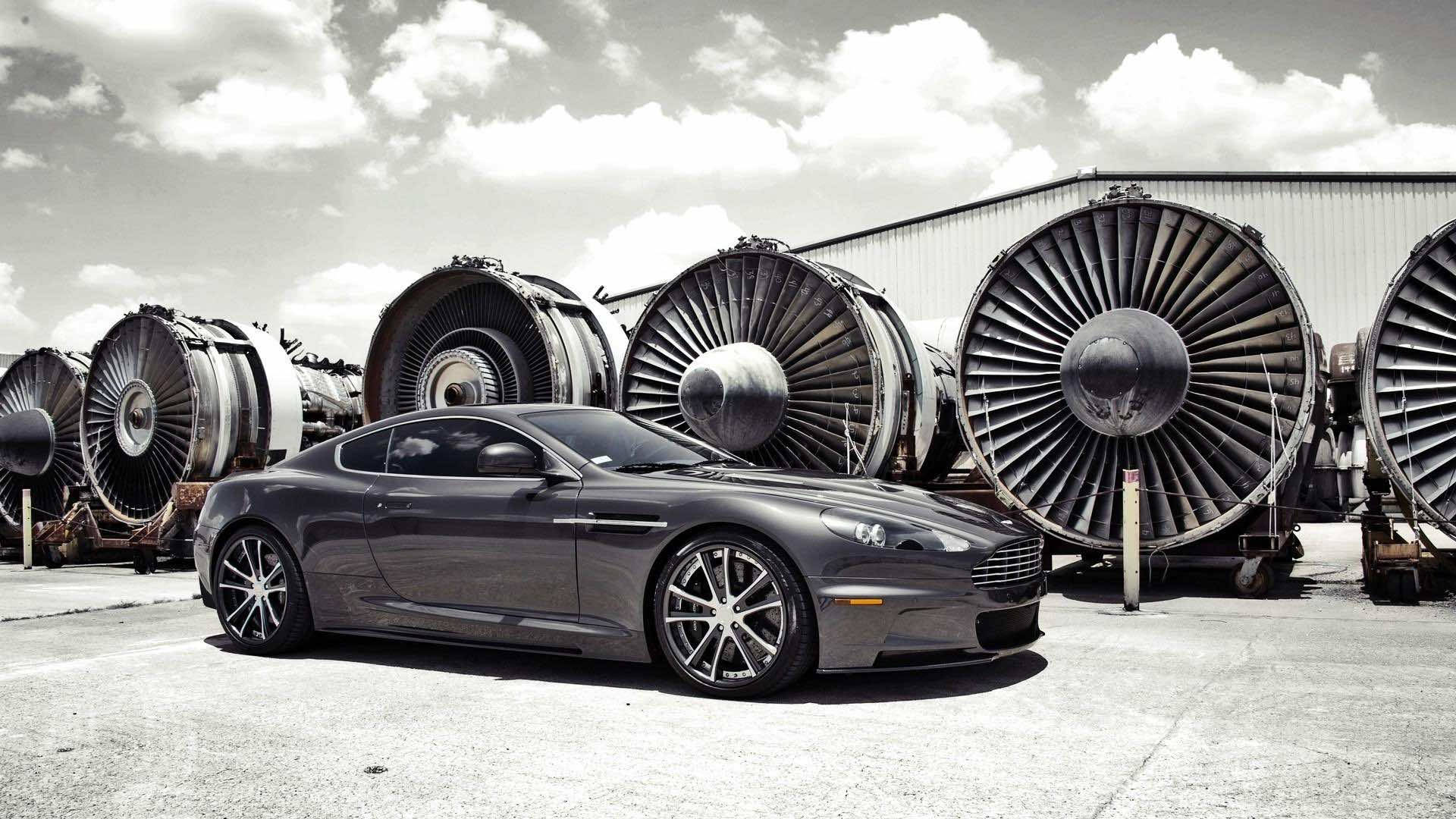 黑白 桌布 壁紙 — 一架時尚的灰色 Aston Martin 跑車停放在巨大噴射引擎前，天空多雲。