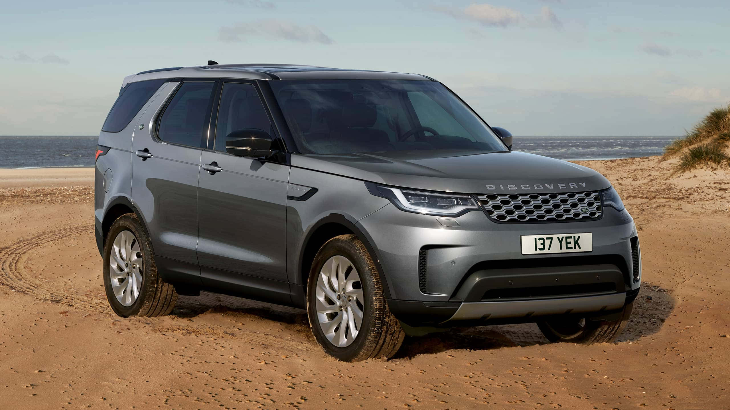 Land Rover Discovery 桌布 壁紙 — 一架時尚的灰色 Land Rover Discovery SUV 停泊在沙灘上，背景是海洋和沙丘。