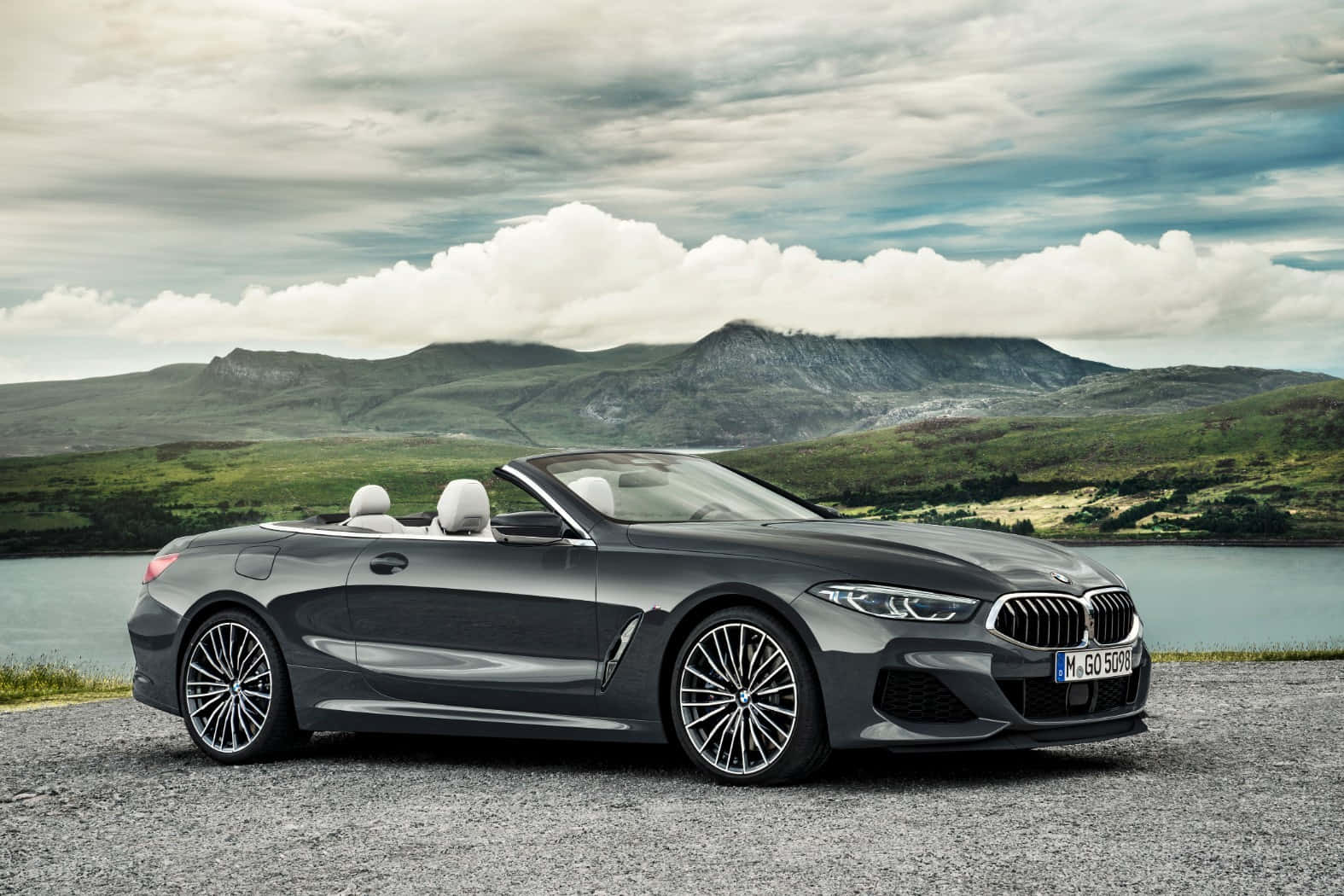 Bmw 8 Series 桌布 壁紙 — 一架時尚的灰色BMW敞篷車停泊在湖邊的礫石岸上，背景是起伏的綠色山丘和多雲的天空。