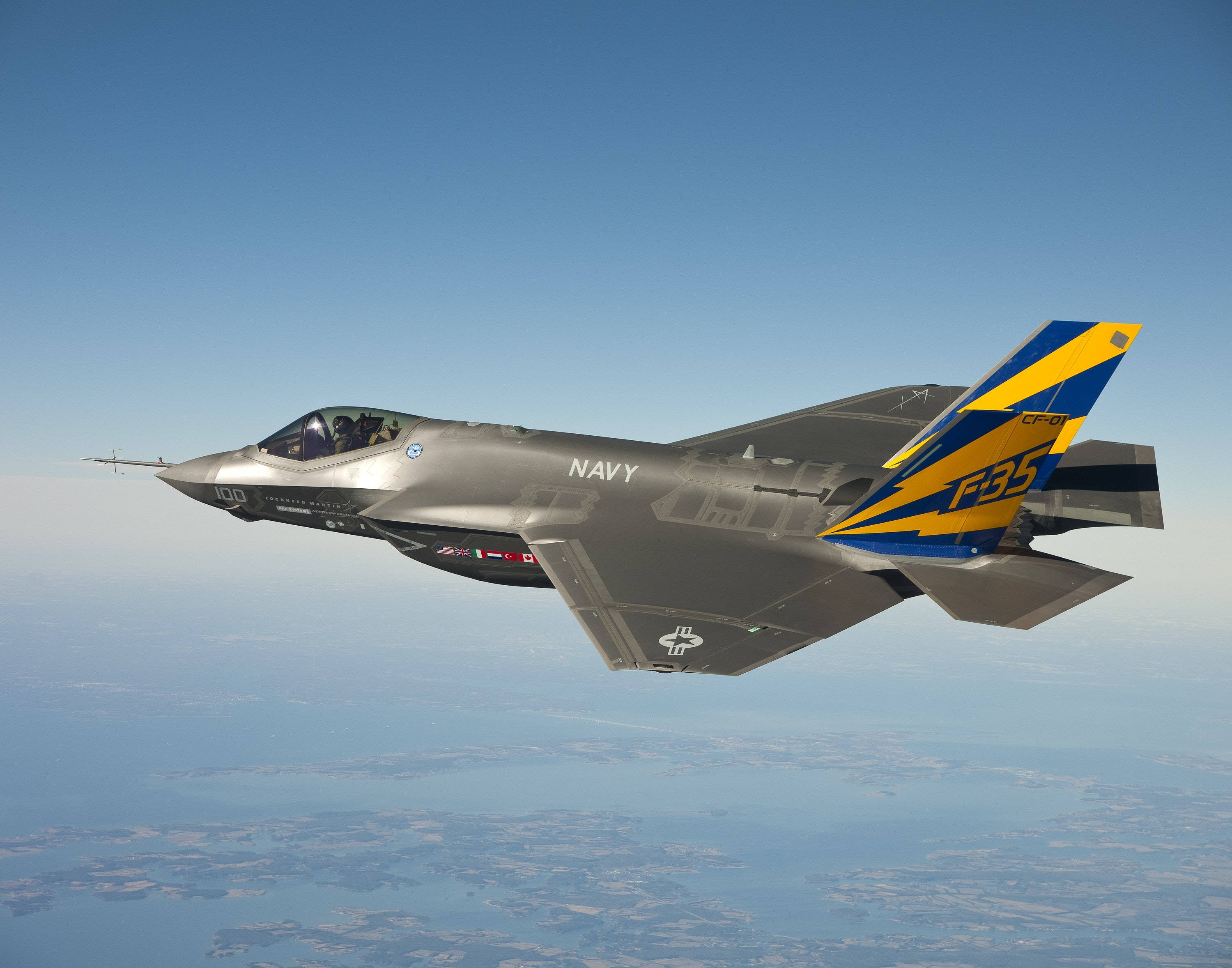 Air Force 桌布 壁紙 — 一架時尚的灰色F35戰鬥機帶有藍黃色尾翼標誌，在清澈藍天下翱翔於沿海景觀之上。