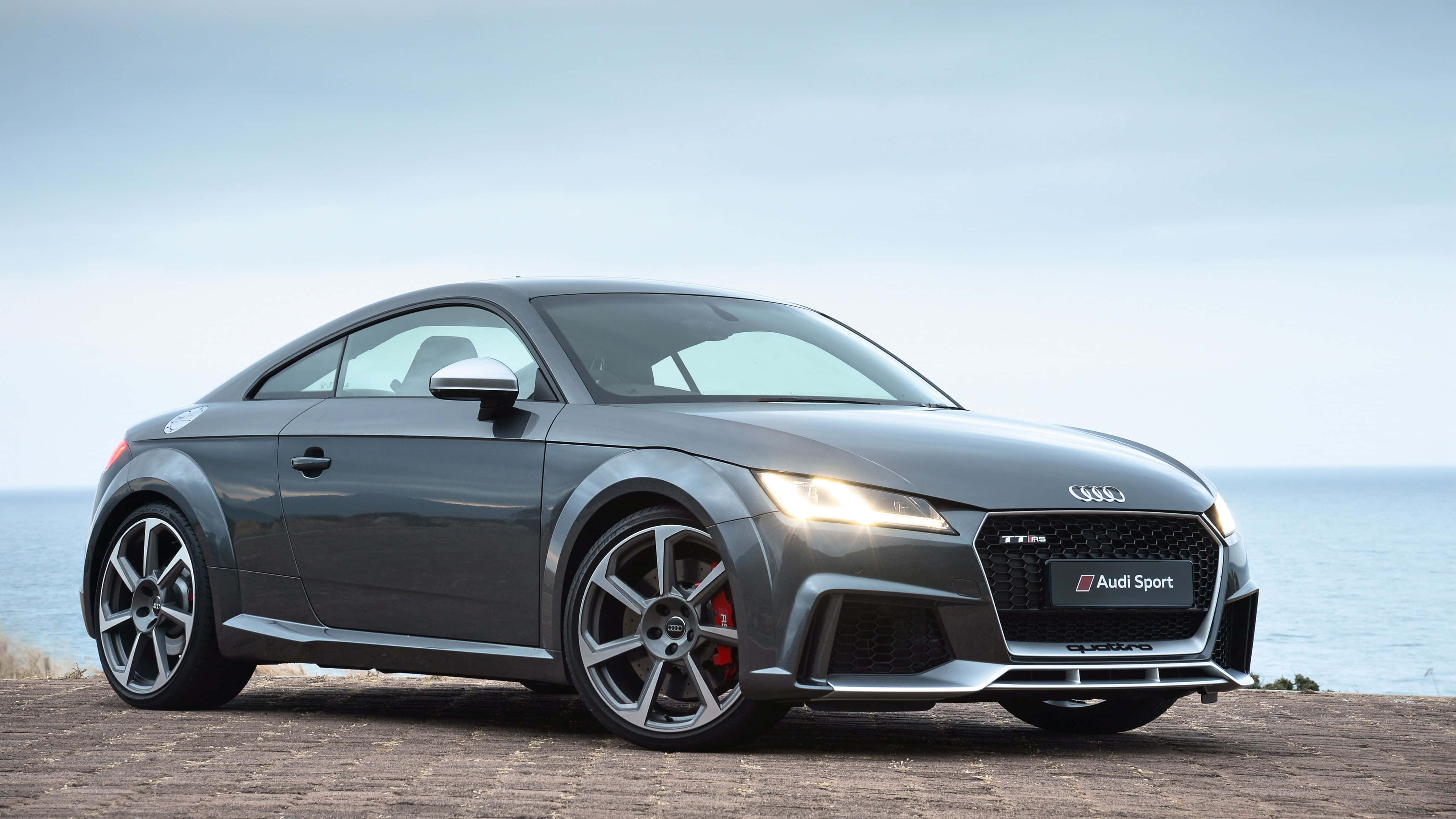 Audi Tt 桌布 壁紙 — 一架時尚的灰色奧迪 TT RS 跑車停泊在懸崖上，俯瞰海洋，車頭燈亮起。