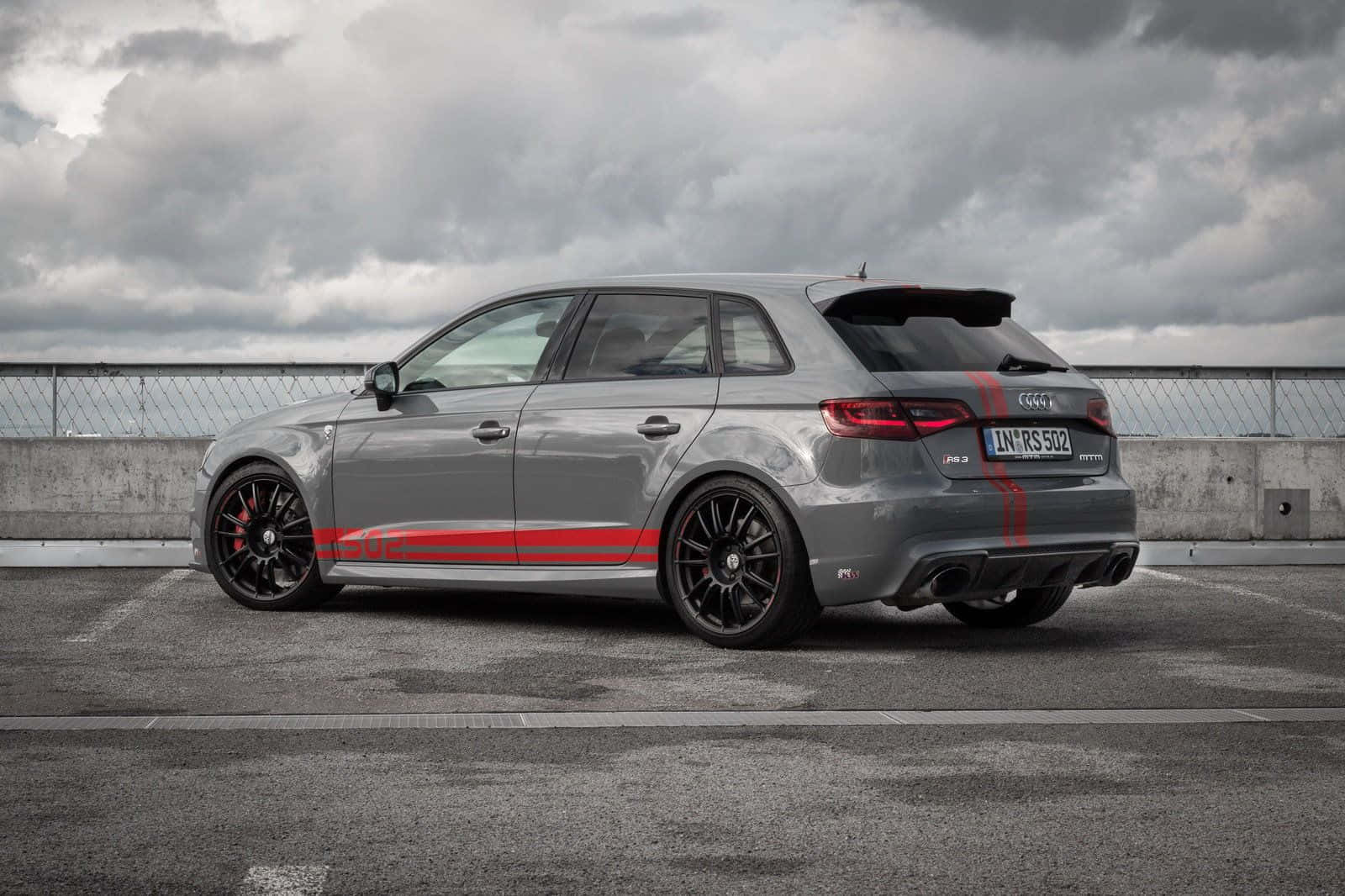 Sedan 桌布 壁紙 — 一架時尚的灰色奧迪RS3 Sportback，帶有鮮明的紅色條紋，停泊在混凝土天橋上，背景是戲劇性的多雲天空。