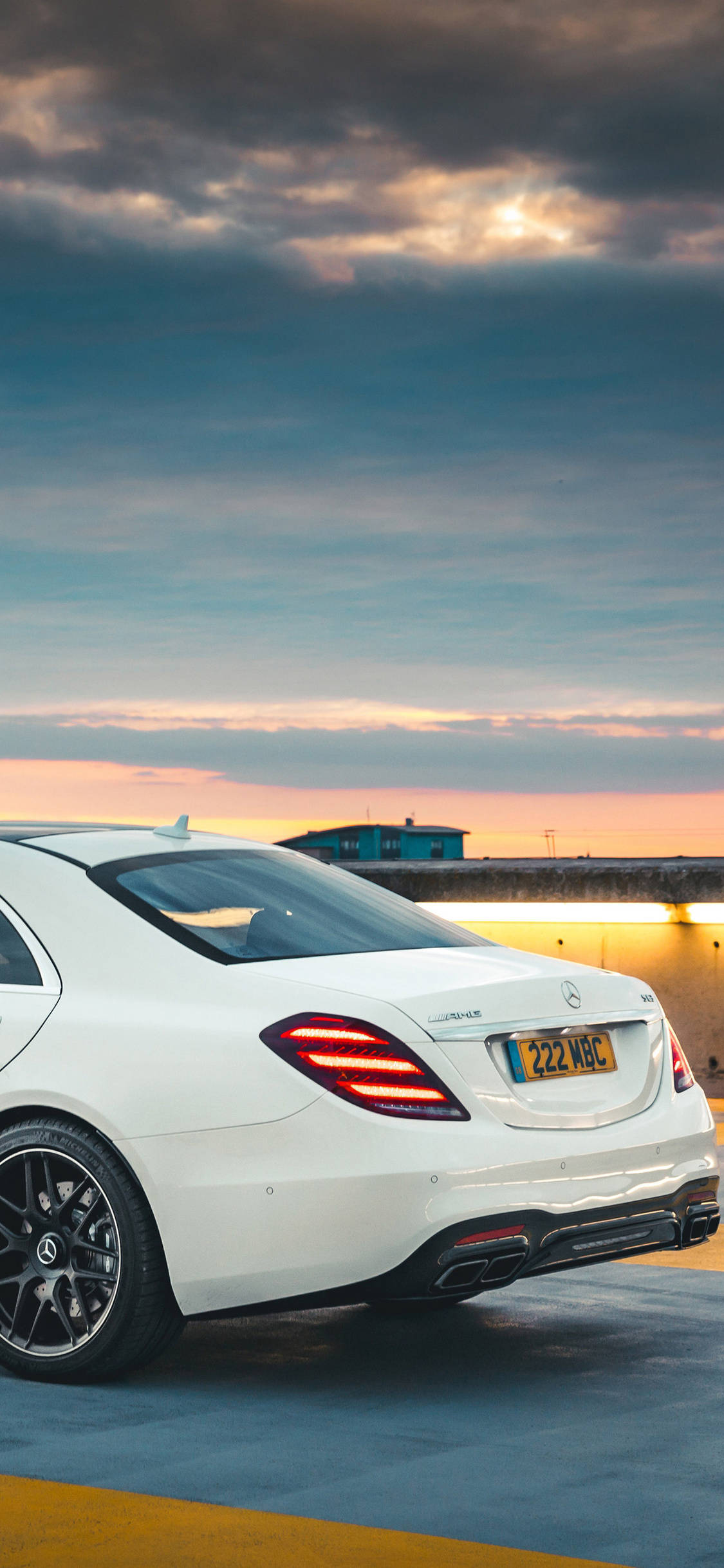 賓士 iPhone 桌布 壁紙 — 一架時尚的白色 Mercedes-Benz 跑車在日落時停泊，帶有發光的尾燈和戲劇性的多雲天空。