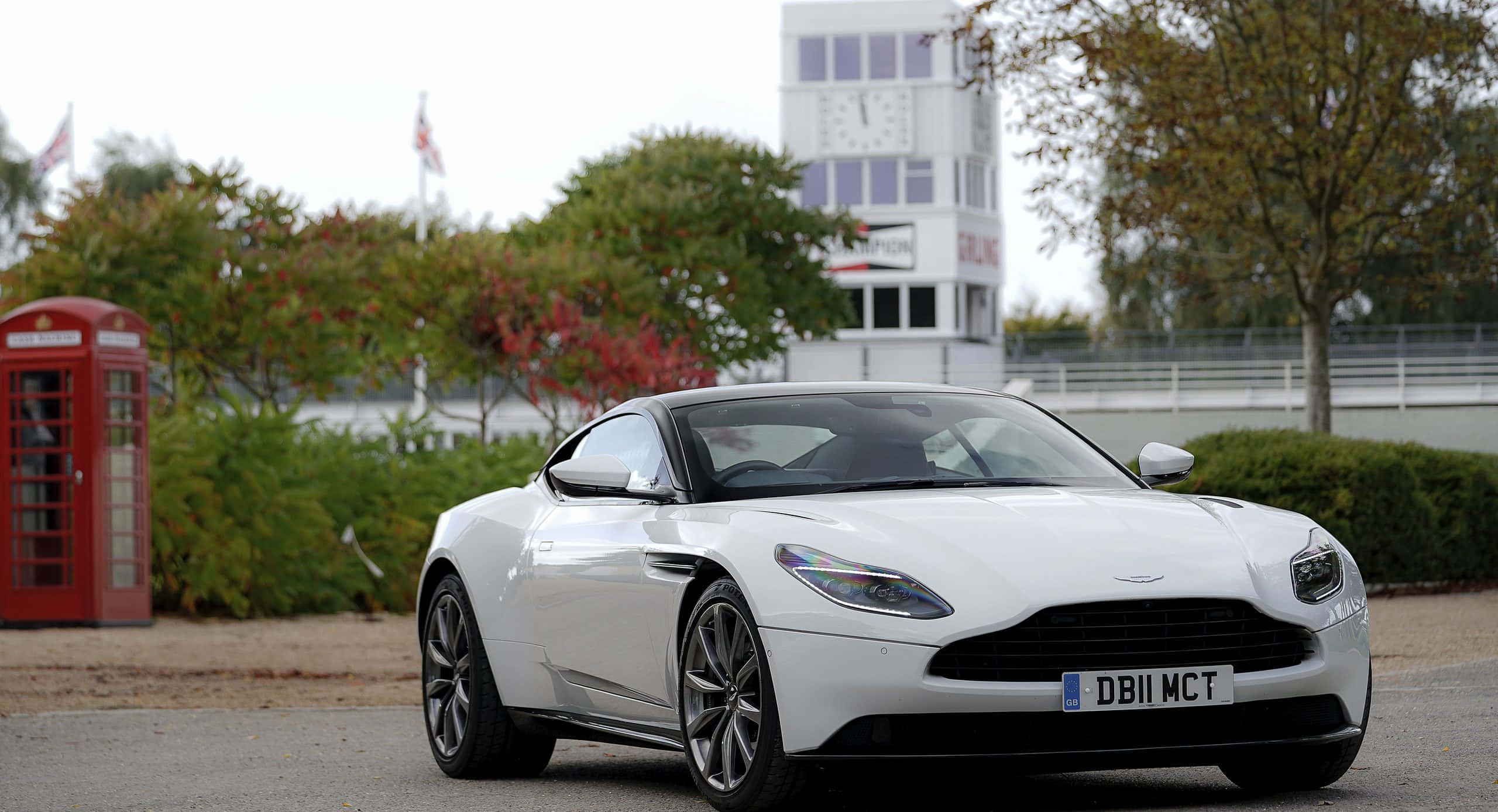 Aston Martin Db11 桌布 壁紙 — 一架時尚的白色Aston Martin DB11停放在經典的紅色英國電話亭旁，背景有秋樹和鐘樓。