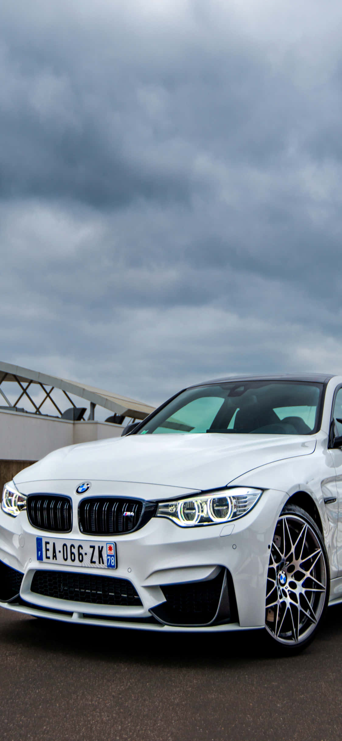 iPhone X Bmw 桌布 壁紙 — 一架時尚的白色BMW M4跑車停放在戲劇性的多雲天空下，背景是現代建築。