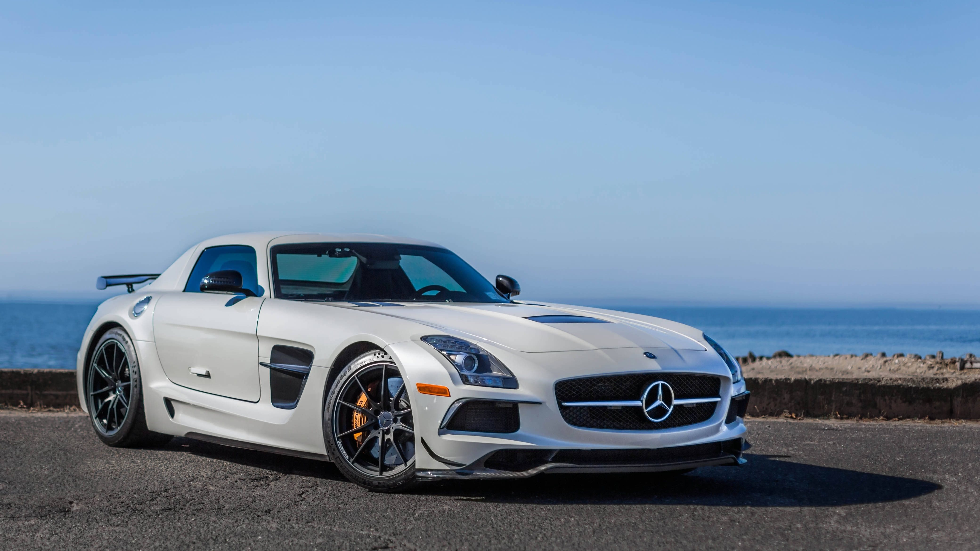 amg iPhone 桌布 壁紙 — 一架時尚的白色Mercedes SLS AMG跑車停泊在海濱道路上，背景是海洋和清澈的藍天。