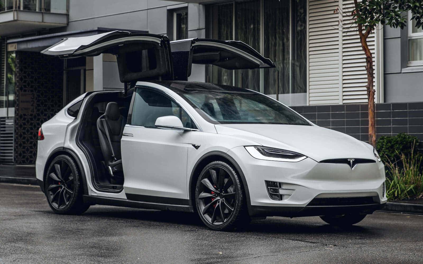 特斯拉 桌布 壁紙 — 一架時尚的白色Tesla Model X，鷹翼門打開，停泊在濕潤的都市街道旁，旁邊是現代建築。