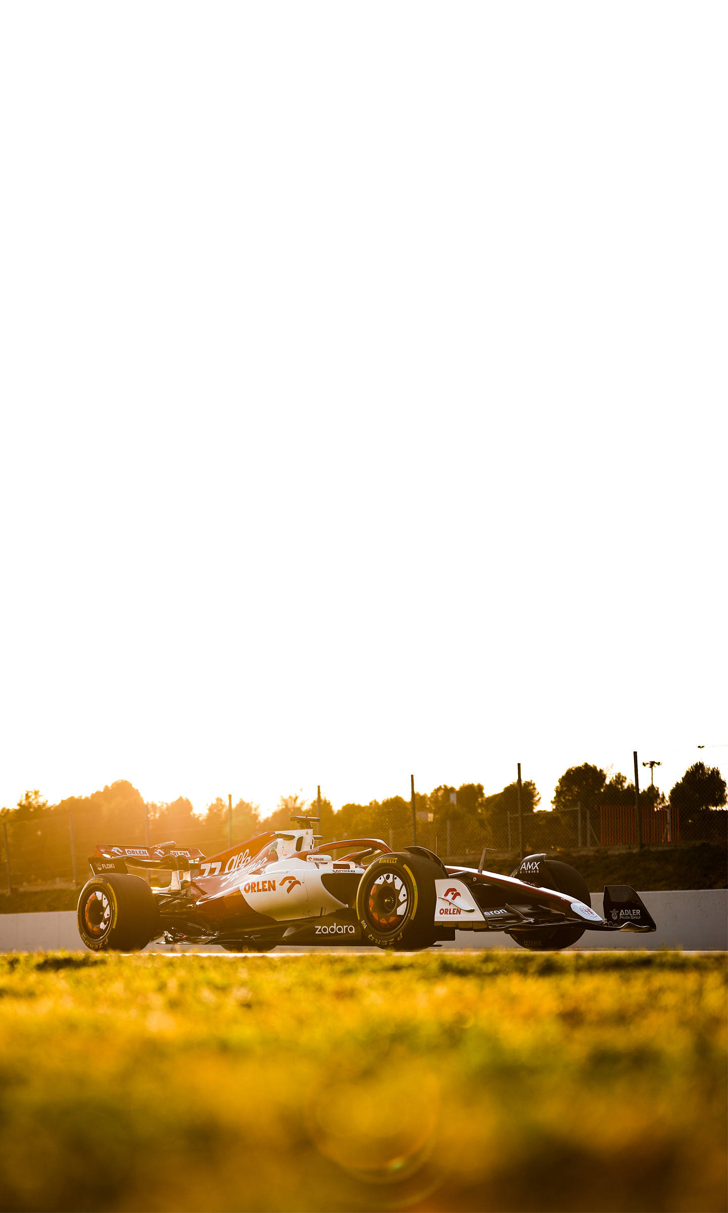 F1 iPhone 壁紙 桌布 壁紙 — 一架時尚的白色和橙色賽車在夕陽下沿著賽道滑行，前景是金色的草地。