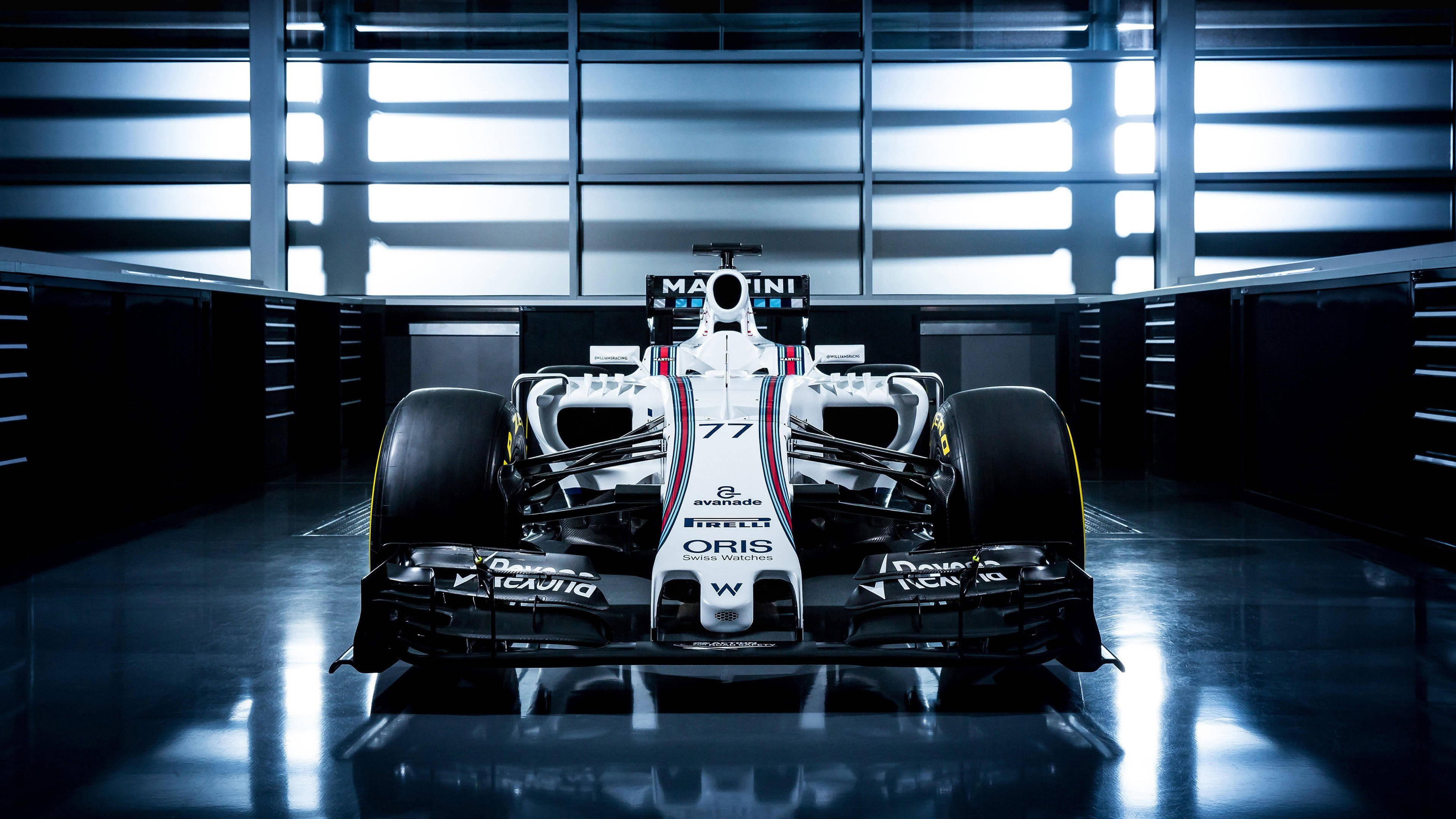 F1 桌布 壁紙 — 一架時尚的白色和黑色一級方程式賽車，編號77，位於高科技車庫中央，上方有明亮的燈光。