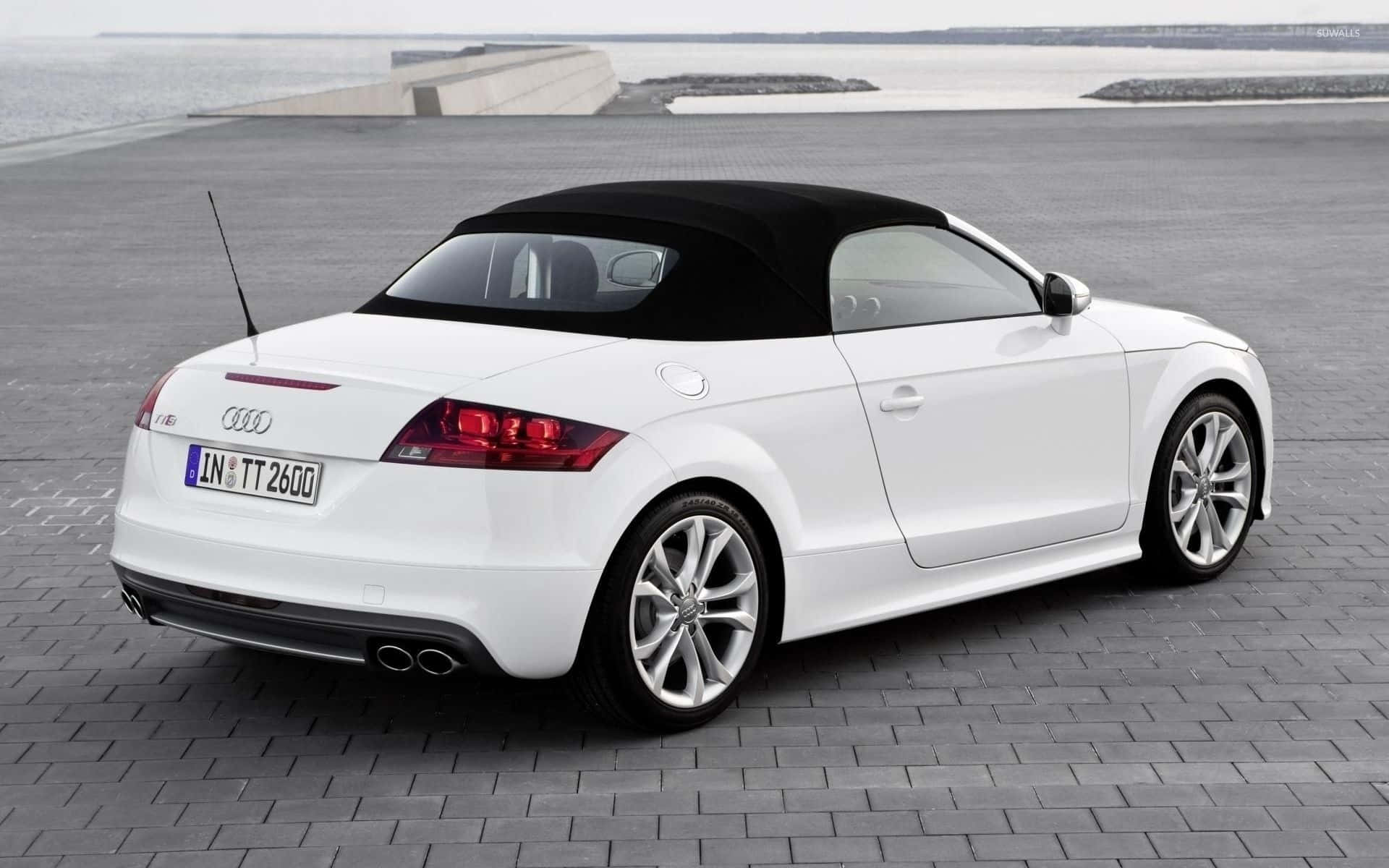 Audi Tt 桌布 壁紙 — 一架時尚的白色奧迪TT Roadster，配備黑色軟頂，停泊在寧靜海濱旁的灰色磚砌廣場上。