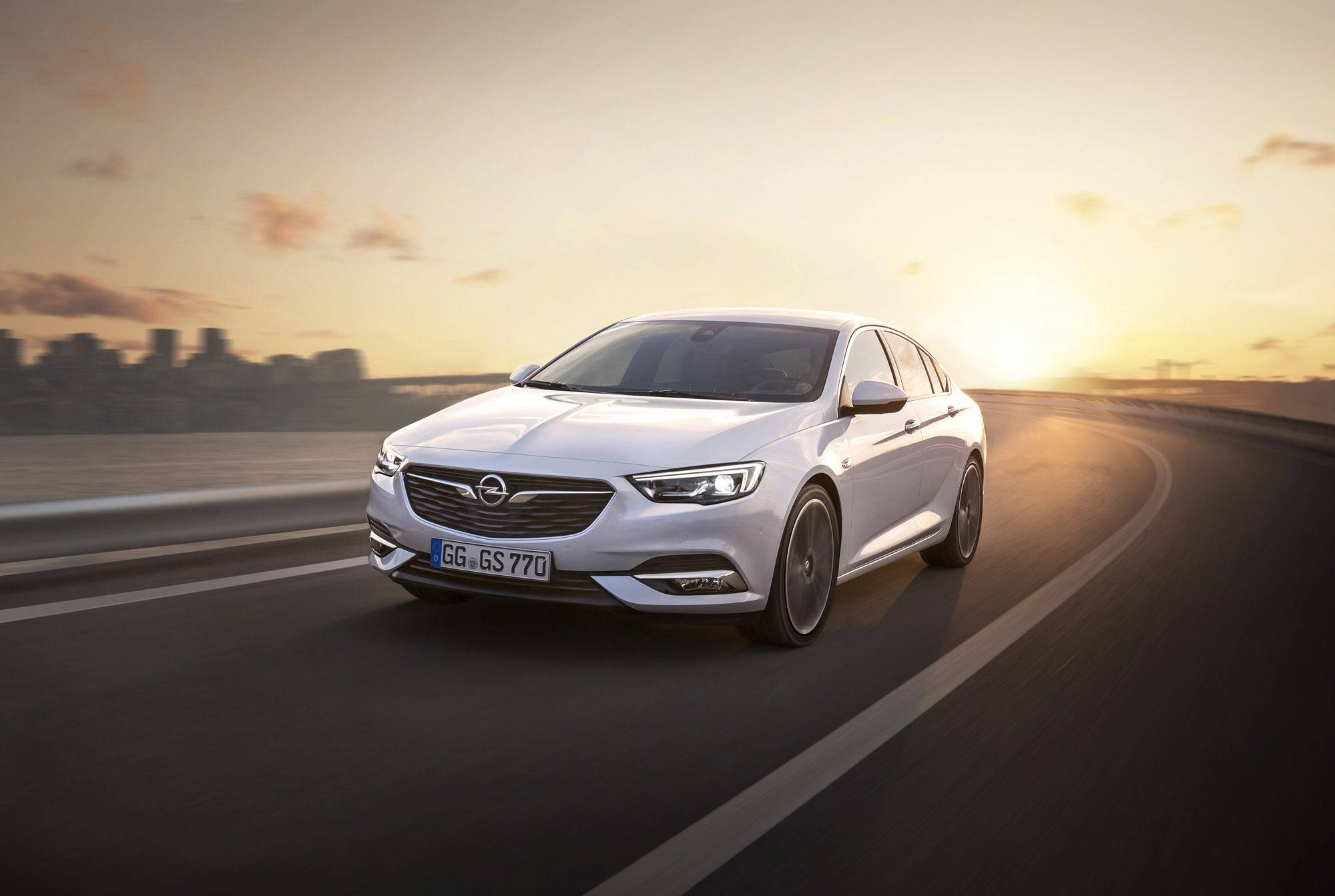 Opel 桌布 壁紙 — 一架時尚的白色歐寶Insignia GS在日落時分沿著彎曲的高速公路行駛，遠處是模糊的城市天際線。