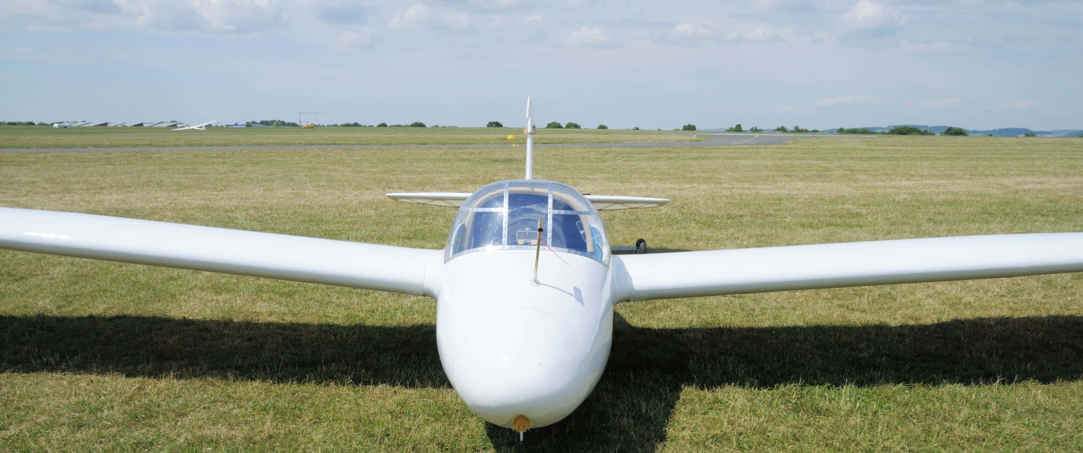 3440x1440p Small Planes 桌布 壁紙 — 一架時尚的白色滑翔機停在陽光明媚的草地上，天空部分多雲。