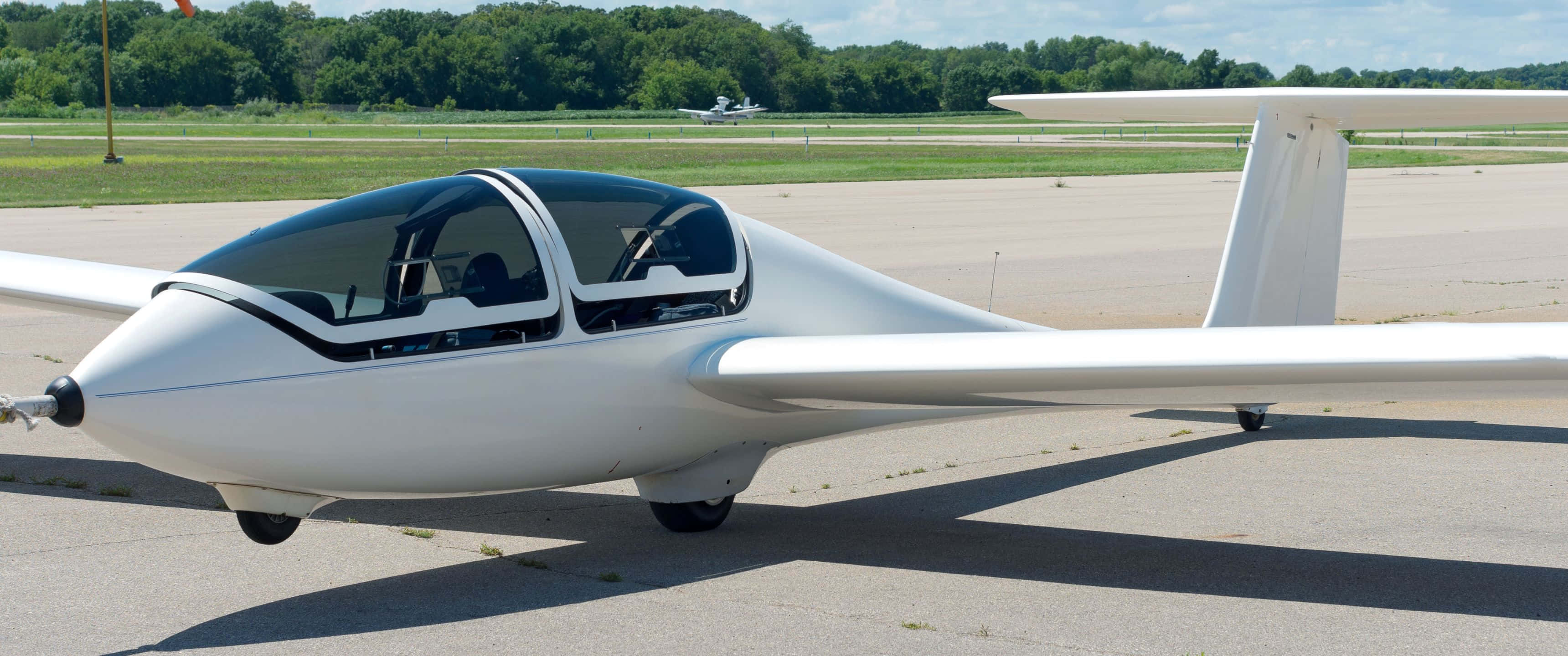 3440x1440p Small Planes 桌布 壁紙 — 一架時尚的白色滑翔機停在陽光明媚的停機坪上，遠處有樹木和另一架飛機。